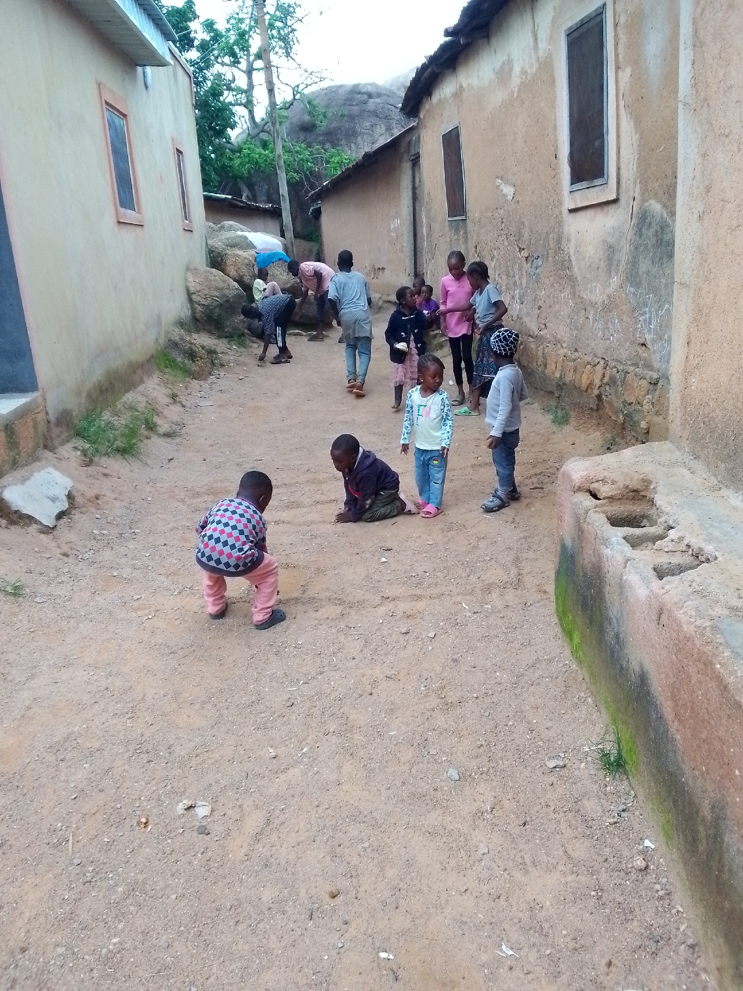 In the city of Unknown, Unknown, a group of children are gathered on a dusty, dirt path between two buildings. One building, on the left, is painted a light tan and has two windows. The other building, on the right, is also tan and appears older with a rougher texture. The path is uneven and strewn with small stones and debris.
In the foreground, two young boys are playing. The boy on the left is wearing a checkered shirt and pink pants, bent over as if playing with something on the ground. The boy on the right is wearing a dark hooded jacket and is kneeling.
Further down the path, more children are visible. A young girl in a light blue shirt and jeans stands facing left. Next to her, a boy in a grey sweater and jeans stands looking forward. Several other children are gathered in the background, some standing and some sitting, with one adult figure walking away. In the background, large rocks and trees can be seen, suggesting the area is near a natural landscape.