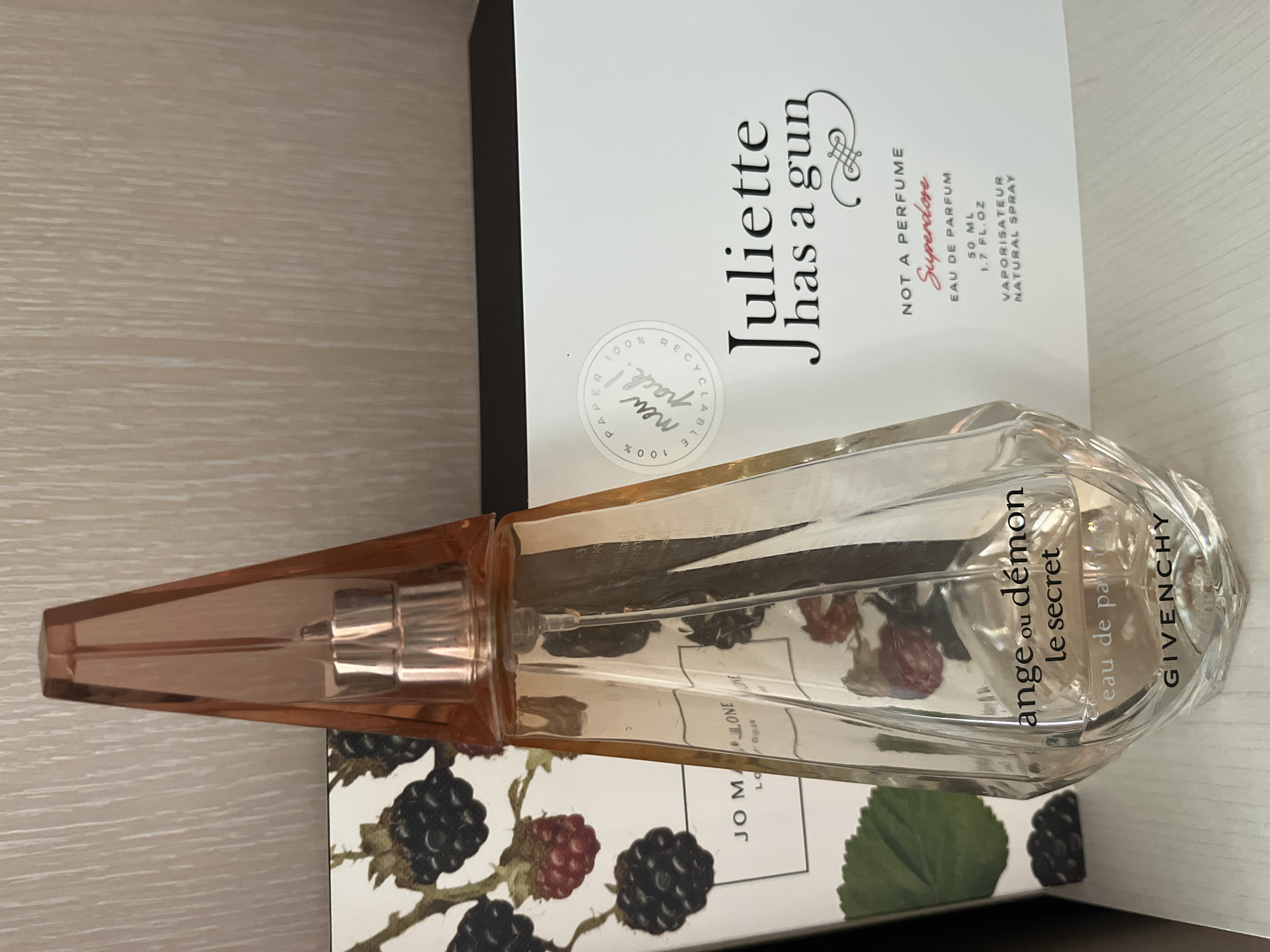 The image shows a still life arrangement of two perfume bottles and boxes. In the foreground, a clear glass perfume bottle with a flared, peachy-colored cap and a faceted base is prominently displayed. The bottle contains a light pink liquid and is labeled with "ange ou démon le secret" and "GIVENCHY" in black text.
To the left of the Givenchy perfume, a portion of a white box with a pattern of blackberries and leaves is visible. The text "JO MALONE LONDON" is partially visible on this box.
To the right of the Givenchy perfume is a white box with black text. It is labeled "Juliette has a gun" and below that, in smaller text, "NOT A PERFUME" and "Superdose". Further down, "EAU DE PARFUM 50 ML 1.7 FL.OZ" and "VAPORISATEUR NATURAL SPRAY" are printed. A circular sticker with the text "100 % PAPER 100 % RECYCLABLE new pack !" is also affixed to this box.
The background consists of a light-colored, textured surface, possibly a wall. The objects are placed on a white surface, likely a shelf or table. The lighting suggests an indoor setting, possibly a display or personal collection.
This image does not provide any information about the city of Komsomolskoye, Ukraine.