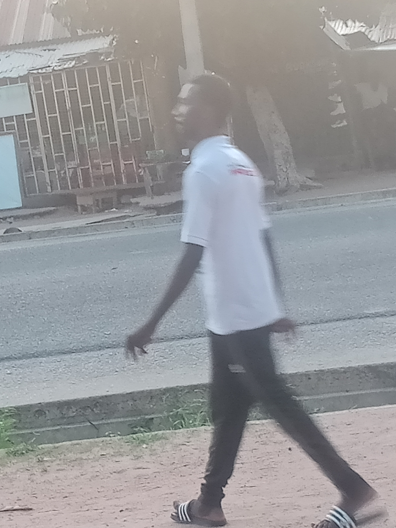 The image shows a person walking on a sidewalk next to a road in Jalingo, Nigeria. The person is wearing a white t-shirt and dark pants, and is carrying a bag. The road appears to be paved, and there are buildings visible in the background. The sidewalk is made of concrete and there is some vegetation growing along the edge of the road. The scene appears to be a typical street scene in a Nigerian city.