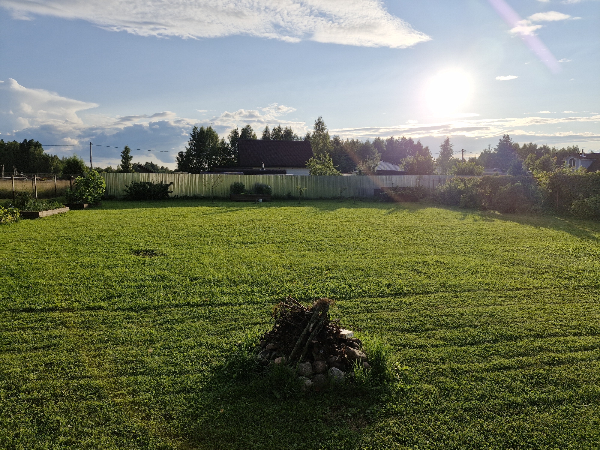This image depicts a sunlit lawn in the village of Sorochkino, Russia. The foreground features a neatly mown green lawn with a small pile of kindling and stones in the center. A metal fence runs across the middle ground, separating the lawn from a wooded area and a few residential buildings. The sky is blue with scattered white clouds, and the sun is bright, casting long shadows across the grass. The overall scene is tranquil and rural, suggestive of a summer evening.