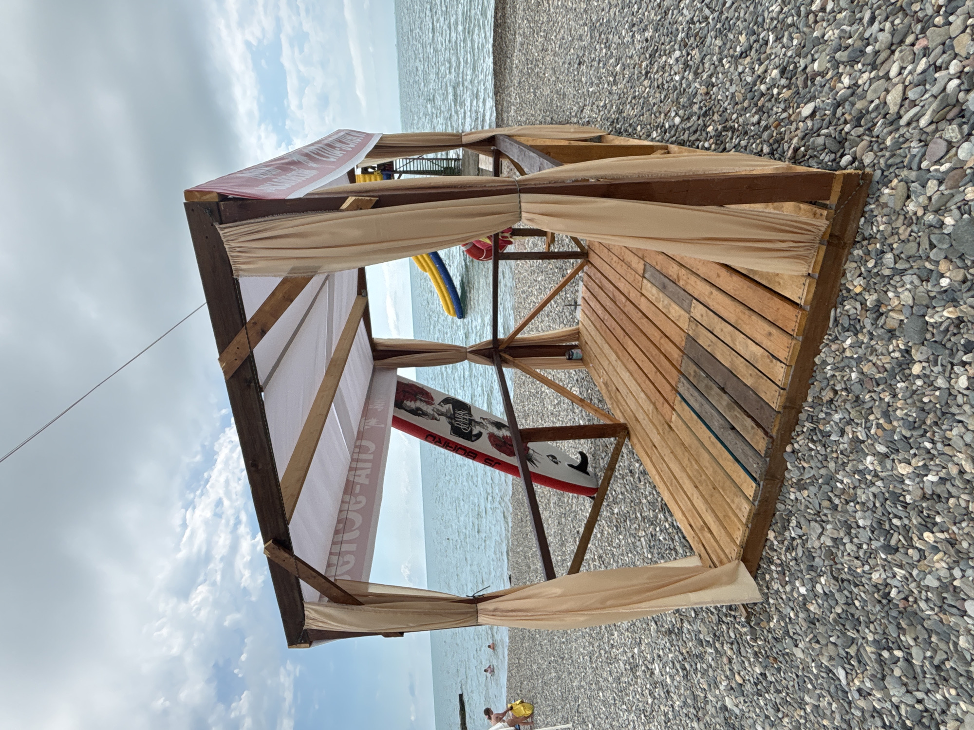 The image shows a wooden gazebo on a pebble beach. The gazebo has a white canopy and light beige curtains. Inside the gazebo, a red and white surfboard with "DARK QUEEN" written on it stands upright. In the background, the sea is visible, with a yellow and blue water slide and a red lifebuoy on a ladder to the right. There are a few people in the water and on the beach. The sky is partly cloudy. This scene is typical of a beach resort area in Sochi, Russia, likely during the summer tourist season.