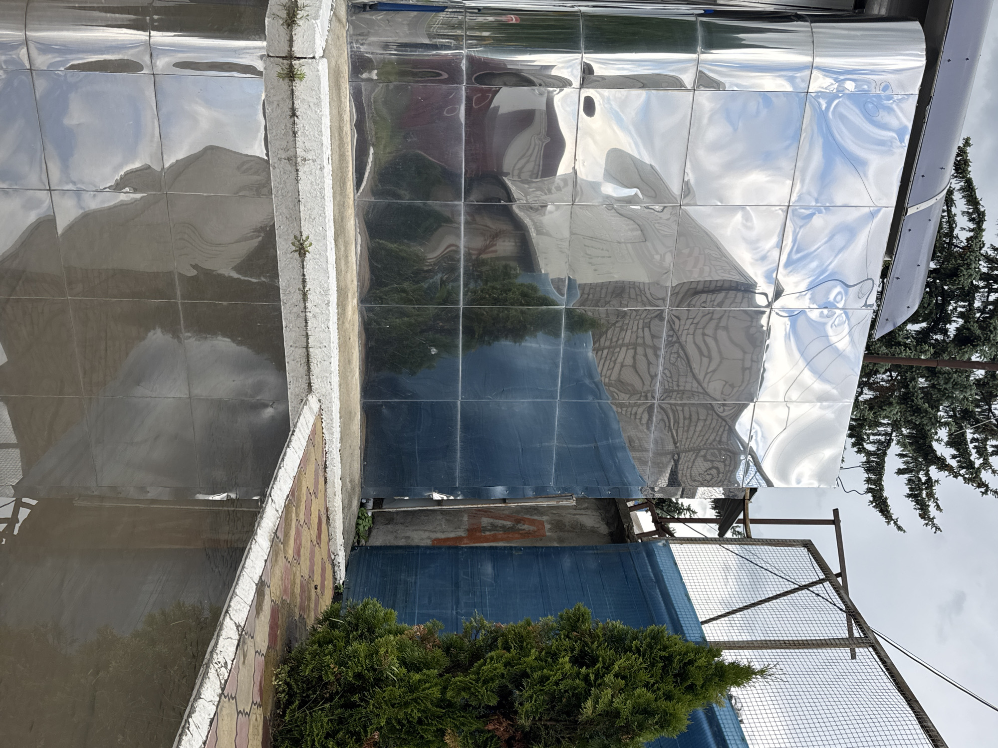 A reflective, metallic, cuboid structure stands in an outdoor environment, possibly in the city of Karachayevsk, Russia. The structure is clad in highly polished, square metal panels that create distorted reflections of the surroundings. These reflections include the sky, clouds, trees, and what appear to be other buildings or structures.
In the background, a fence made of metal mesh and posts separates the area. Adjacent to the metallic structure is a blue, opaque barrier or wall, and behind that, a concrete wall with a large, red, stylized letter "A" painted on it. To the right of these structures is a conifer tree.
The ground in front of the metallic structure is paved with interlocking tiles in red and yellow patterns. A wet surface, possibly a puddle or recently rained-on pavement, reflects the metallic structure and the sky. Small green plants are growing in the crevices of a white border along the base of the metallic structure and the paved area. The overall impression is one of an industrial or utilitarian outdoor space with a striking, modern architectural element.