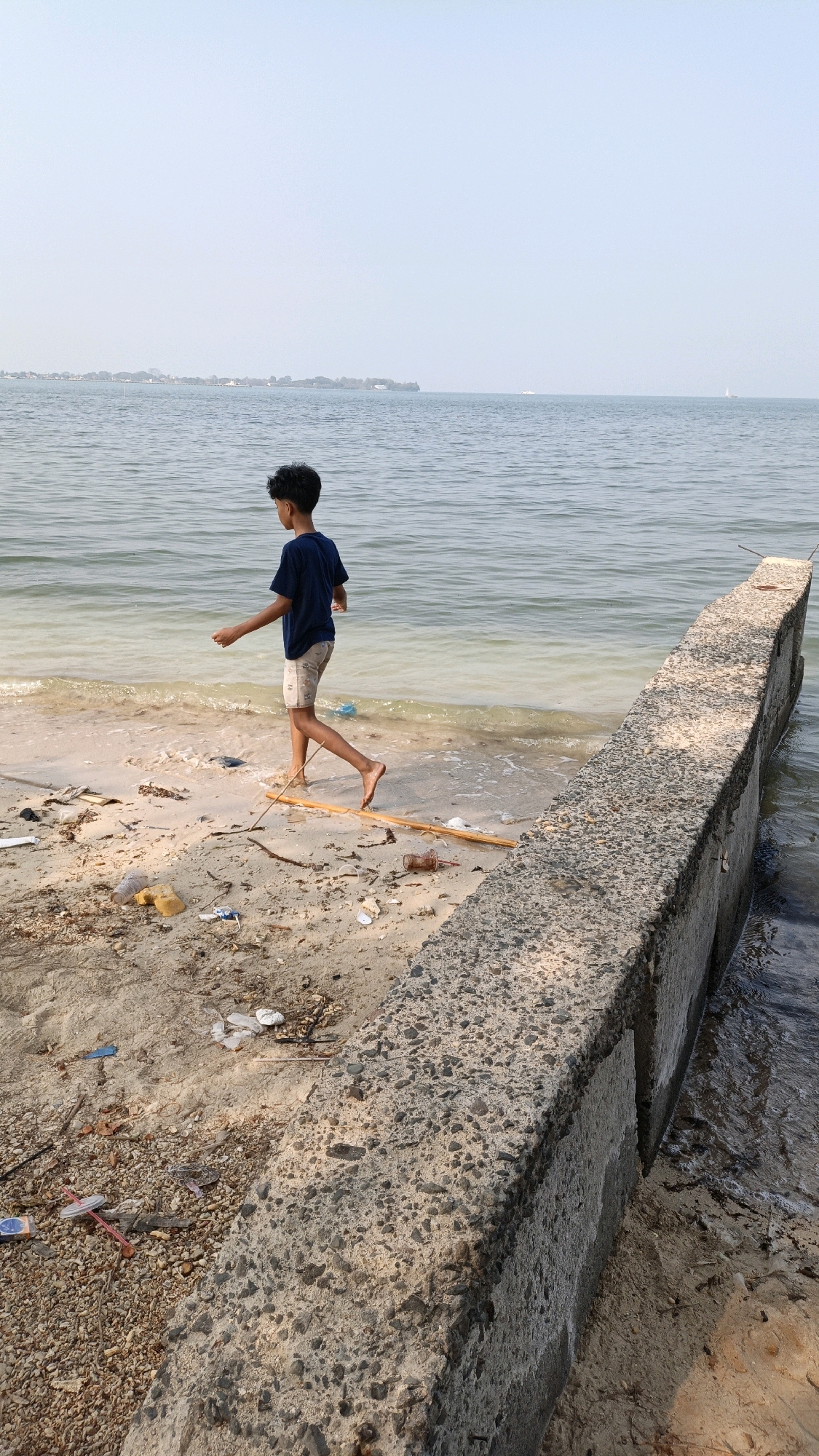 A young boy with dark, curly hair is walking barefoot along a sandy shore. He is wearing a dark blue t-shirt and light-colored shorts. He is facing away from the camera, and his left leg is extended forward. The shore is littered with various pieces of trash, including plastic bottles, wrappers, and other debris. A concrete seawall runs along the right side of the image, separating the beach from the calm water. The water is a murky green color, and the sky above is clear and pale blue. In the distance, across the water, is a faint outline of land with buildings and trees, possibly a city skyline. There are a couple of distant boats visible on the water. The overall scene suggests a somewhat neglected beach environment. Based on the context provided, this location is in Cikal, Indonesia.