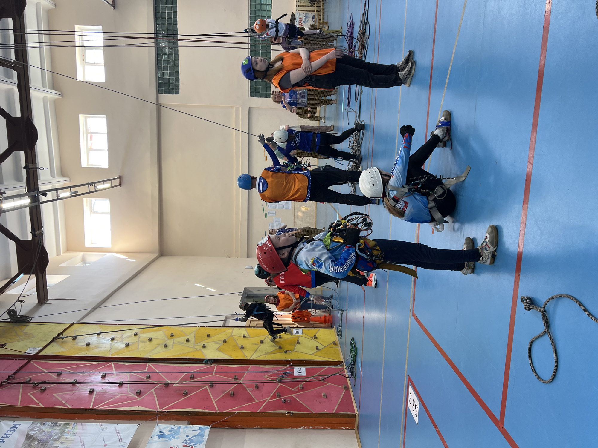 Several people are participating in an indoor rock climbing activity at a climbing wall in Berezniki, Russia. The climbing wall is divided into sections of different colors including red and yellow. Participants are wearing climbing harnesses and helmets, with ropes attached for safety. Some are actively climbing, while others are standing on the floor, likely waiting their turn or assisting. The indoor space has a blue floor marked with red lines. Large windows are visible in the upper part of the wall. Banners hang near the wall’s base.