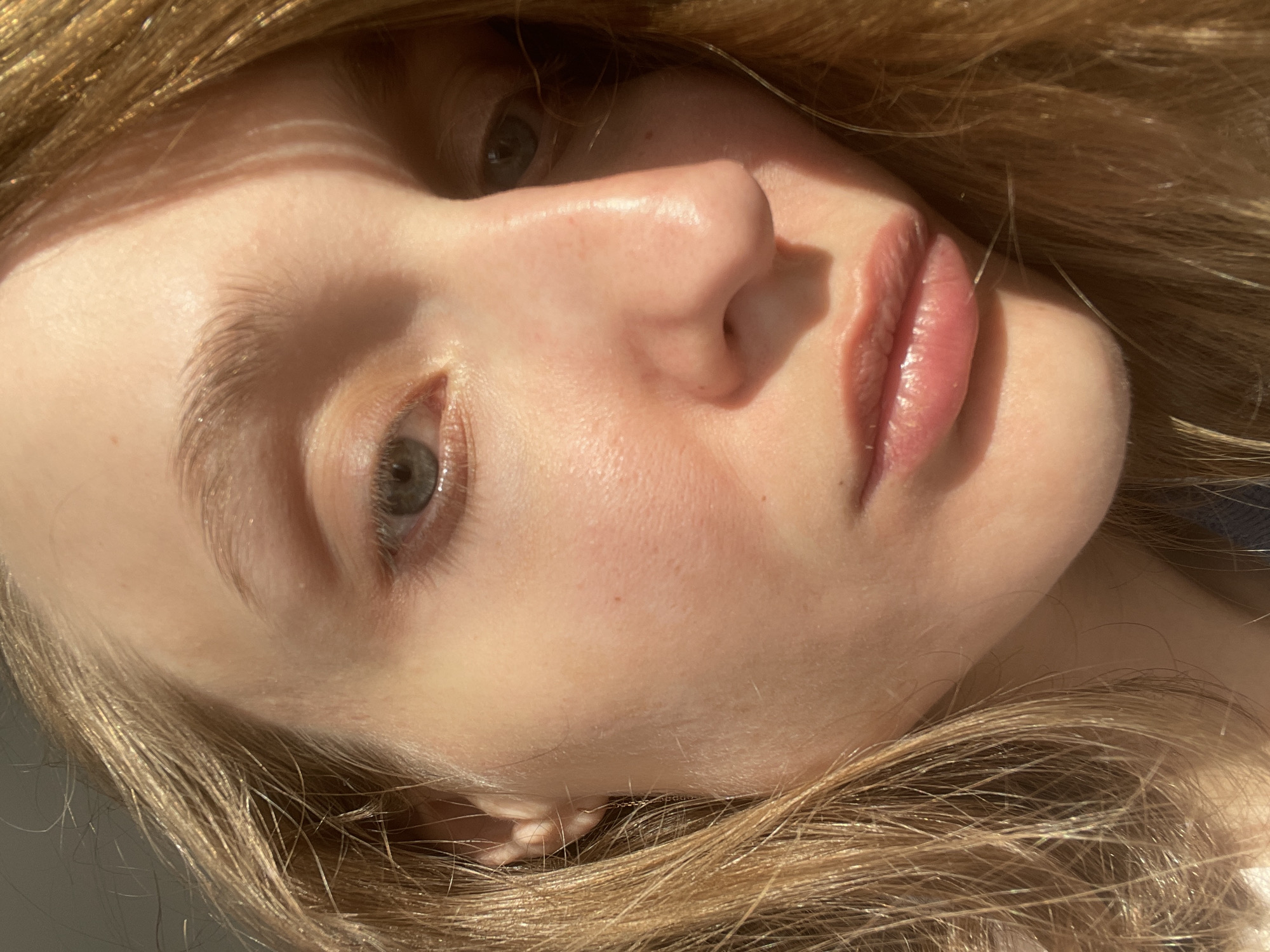 Close-up portrait of a fair-skinned person with light blonde hair, tilted to the side. The person has gray-green eyes, a small nose, and full lips. The skin appears smooth and has a slight sheen, possibly due to lighting or moisturization. The overall scene appears to be indoors with natural lighting, casting shadows across the face. There is no information available from the image or the context provided that indicates the location is Sepych, Russia.