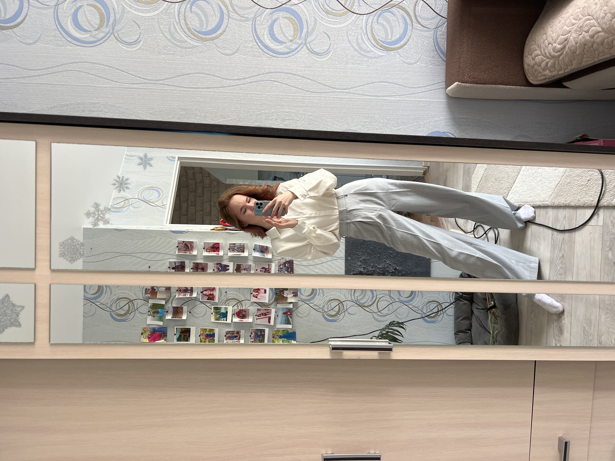 A woman is reflected in a multi-panel mirror in a room. The room has wallpaper with a swirl pattern. The mirror has multiple images attached to the mirror and reflects her image. In the background is a couch.