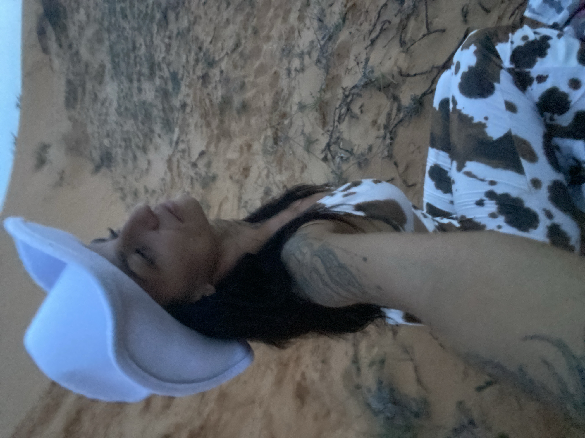 A woman wearing a white cowboy hat and a cow-print outfit is sitting on sand dunes. Her eyes are closed and her head is tilted back as if she is enjoying the sun. She has tattoos on her arm. The sand dunes are a light orange color and there are some dry plants scattered around. The sky is a light blue. The location is the sand dunes of Mui Ne, Vietnam.