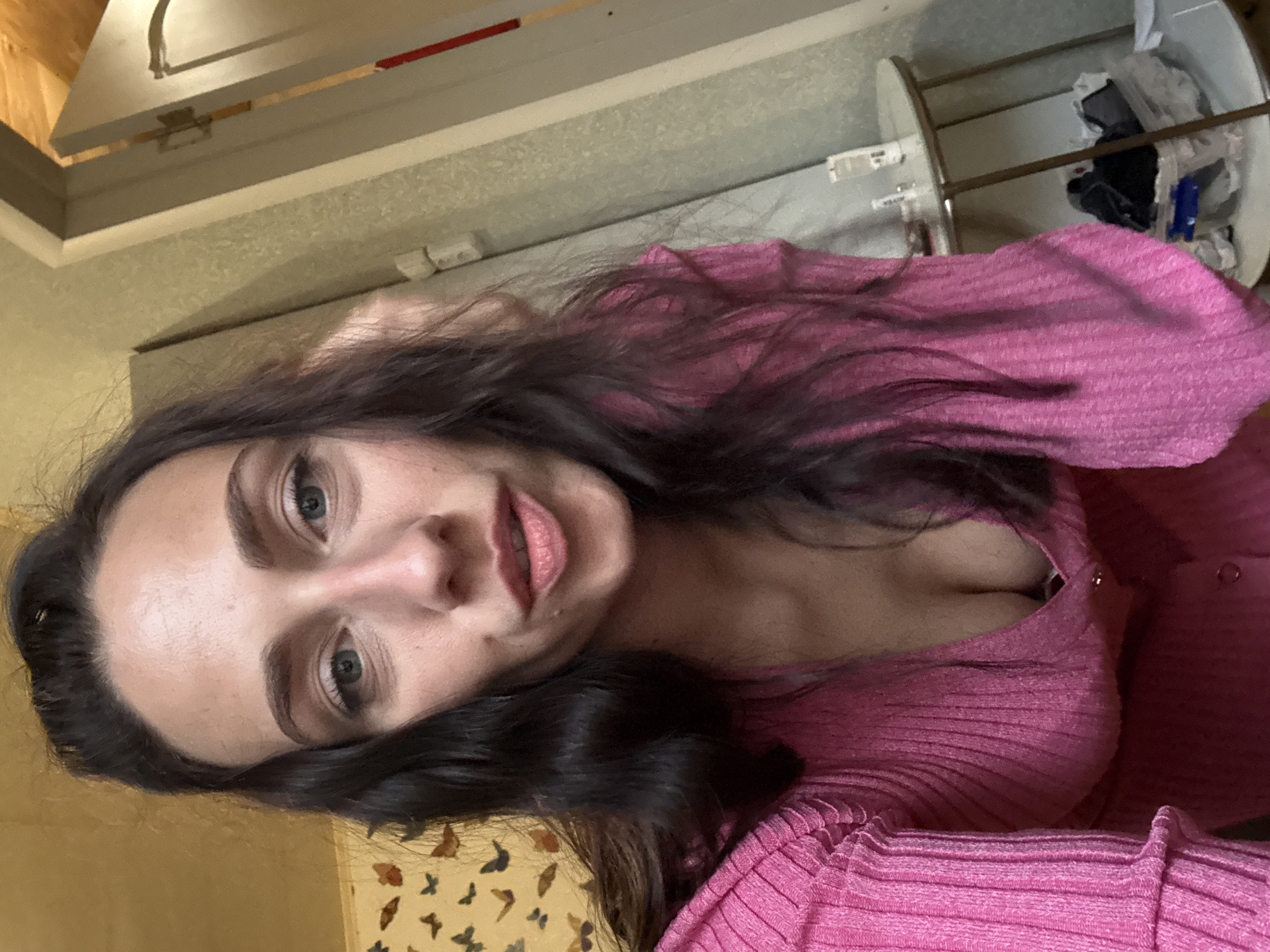 A young woman with long, dark hair and light eyes is taking a selfie in an interior space. She is wearing a pink, ribbed, low-cut shirt. Behind her are neutral-toned walls and a shelf holding various items. The location is not identifiable as Saint Petersburg, Russia.