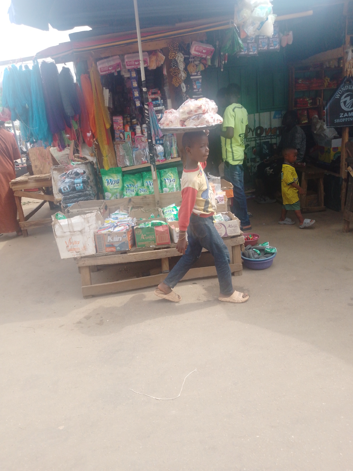 Here is a detailed description of the image:
The image shows a young boy balancing a tray of what appears to be packaged food items on his head, possibly in a market setting. He is wearing a long-sleeved shirt, jeans, and sandals, walking on a light-colored pavement. Behind him is a bustling scene with several people, including a child in a yellow and green striped shirt. Stalls are visible with various goods on display such as colorful fabrics, household products, and toiletries. The stalls are covered by awnings or roofs. In the background, there's a sign that appears to say "ZAM SHOPPING". The image captures a candid moment of everyday life in an urban market area.