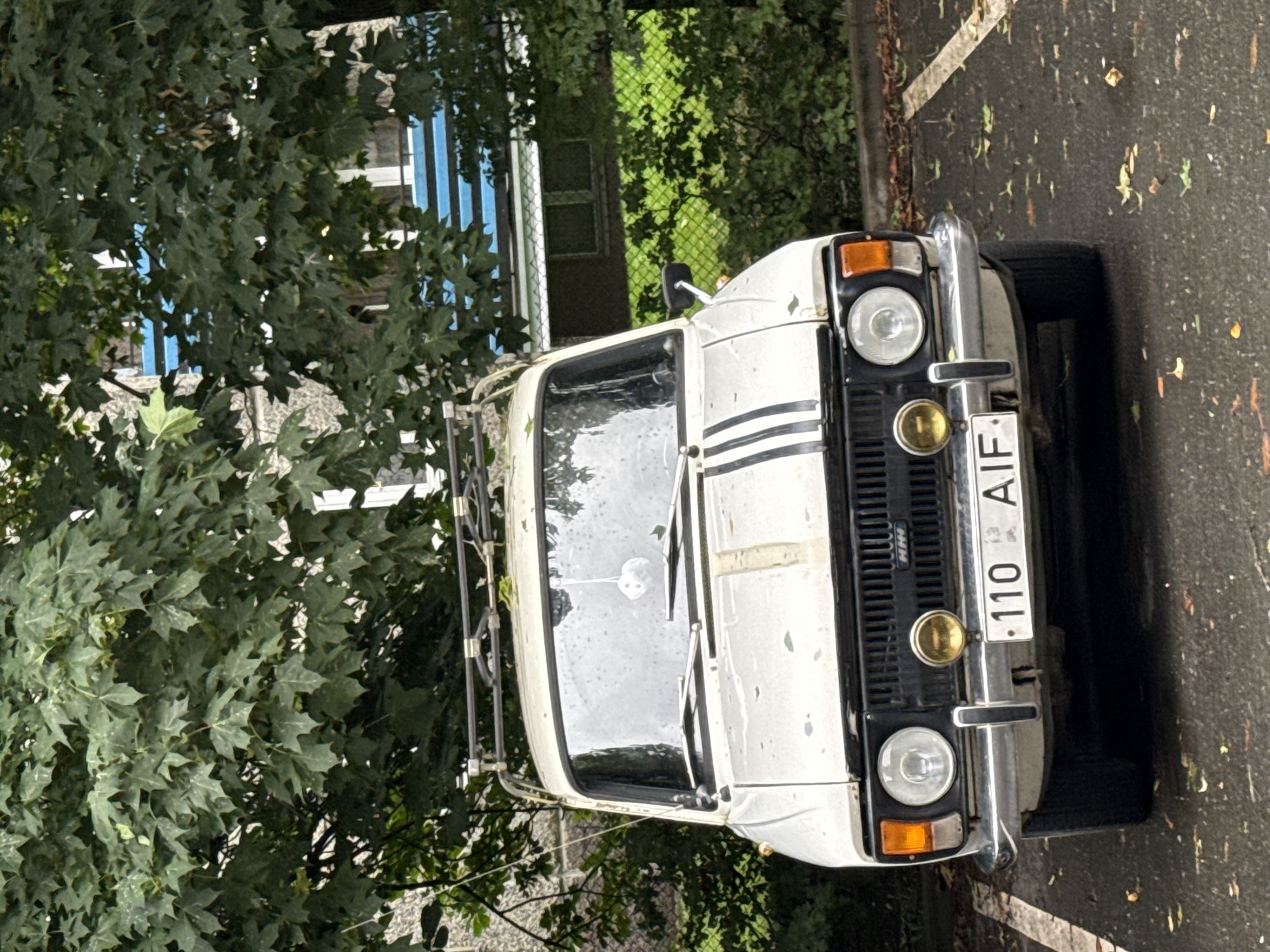 The image shows the front view of a white Moskvitch 2140 parked in a parking spot in Tallinn, Estonia. The car has a roof rack and two black stripes running down the hood. It also has two round headlights, a small yellow fog light, and an orange turn signal on each side. The license plate reads "110 AIF". The car is parked under a leafy green tree, and in the background, there is a building with blue horizontal accents. The ground is wet and appears to be asphalt, with some fallen leaves scattered around. The overall scene suggests an overcast day.