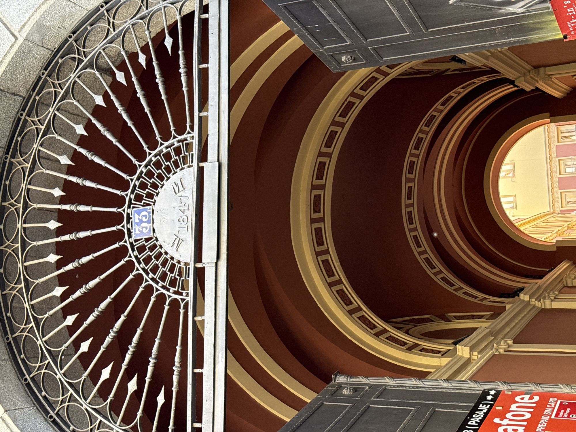 This image is a low-angle shot of an elaborate iron gate with the number "33" on a blue tile and the year "1847" displayed, positioned next to an interior view featuring a series of concentric arched ceilings in a red and beige color scheme. The image composition is unusual, as the gate is oriented vertically on the left side of the frame, while the architectural interior is seen above and to the right.
The gate is ornate with a radial design, and the "33" suggests a street address, while "1847" might indicate the year of the building's construction or a significant historical event. The concentric arches of the building’s interior create a tunnel-like effect leading towards a bright opening in the distance. The architecture features decorative patterns and rectangular designs. At the bottom right, there is a partial view of a sign or advertisement, partially obscured, with the word "afone" visible.
Based on the architectural style and the inscription on the gate, it is likely that the location is Madrid, Spain. This is inferred due to the architectural design being consistent with historical buildings often found in European cities like Madrid.