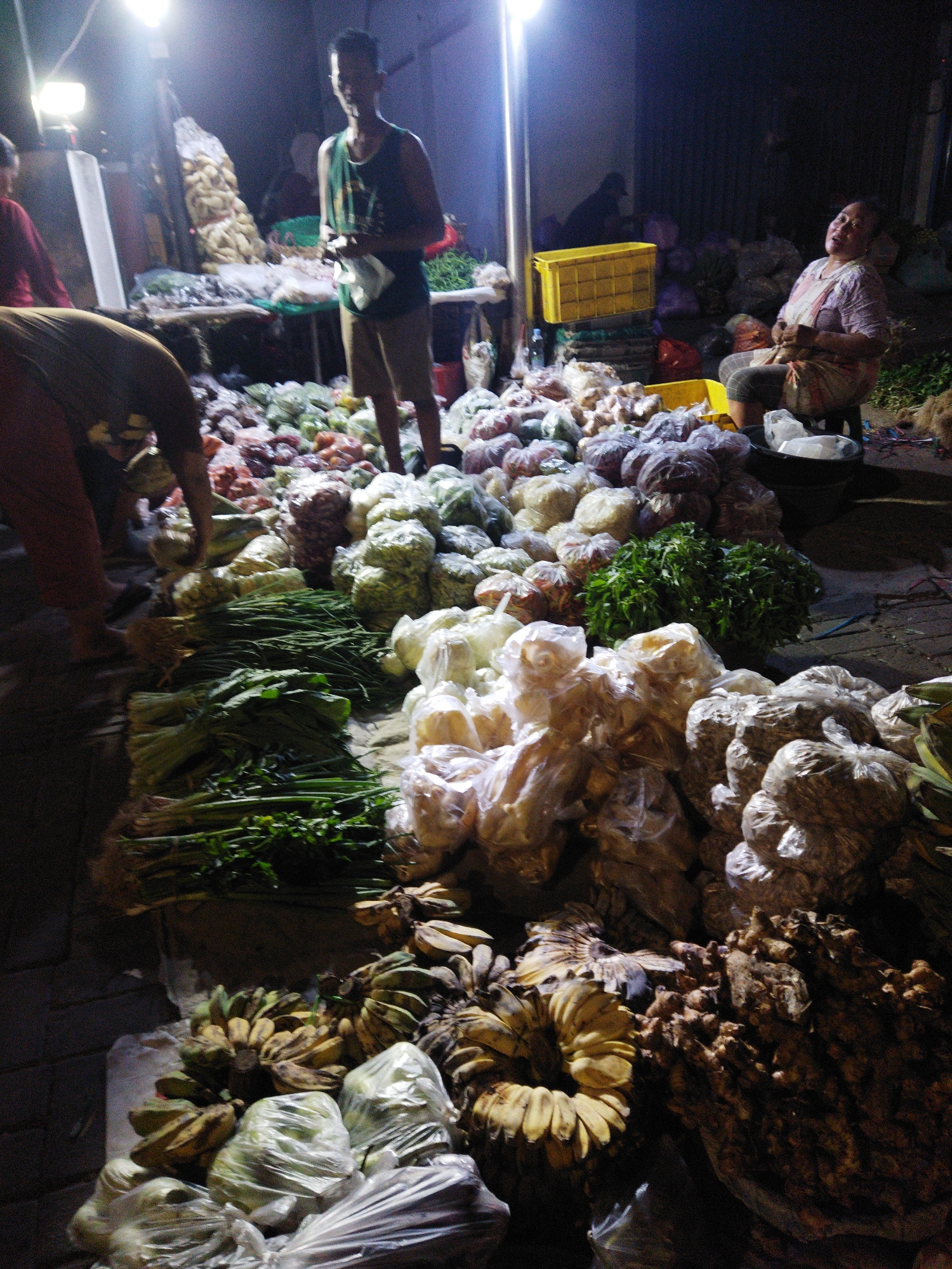 This is a night photograph of an outdoor market stall, likely in Semarang, Indonesia, judging by the presence of typical Indonesian produce and market activities. The scene is dimly lit by artificial lights, creating strong shadows and a somewhat grainy appearance.
In the foreground, a variety of vegetables and fruits are displayed, some wrapped in plastic bags, others loose. Bunches of green onions and leafy greens are prominent, alongside piles of what appear to be ginger or turmeric roots, and several bunches of bananas. Other produce, possibly onions and chilies, are visible in plastic bags.
A man wearing a dark tank top and light-colored shorts stands in the middle ground, holding a plastic bag. He is looking towards the camera. To the right, a woman sits on a stool, wearing a patterned blouse and striped pants, her mouth open as if speaking or laughing. She is looking to her left.
In the background, more produce is visible, including large sacks of what could be corn or dried goods. The overall impression is that of a bustling, albeit dimly lit, night market. The ground is uneven, suggesting it might be a paved street or an area with packed earth. The atmosphere is one of commerce and daily life in a Indonesian urban setting.