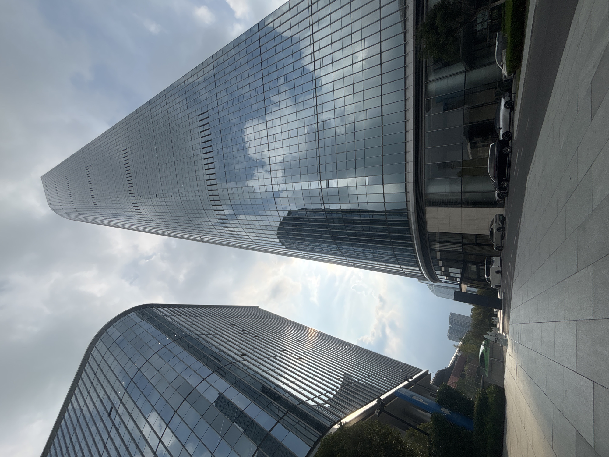 The image shows a low-angle view of modern glass skyscrapers in Suzhou Industrial Park, China. The sky above is partly cloudy with sunlight peeking through. In the foreground, a paved walkway leads towards the buildings, with several cars parked on the street alongside a tree-lined median. One of the buildings on the left has a curved glass facade, while the taller building on the right is a tapering glass skyscraper. The reflections of clouds and sky are visible on the glass surfaces of the buildings. There are no people clearly visible in the image.