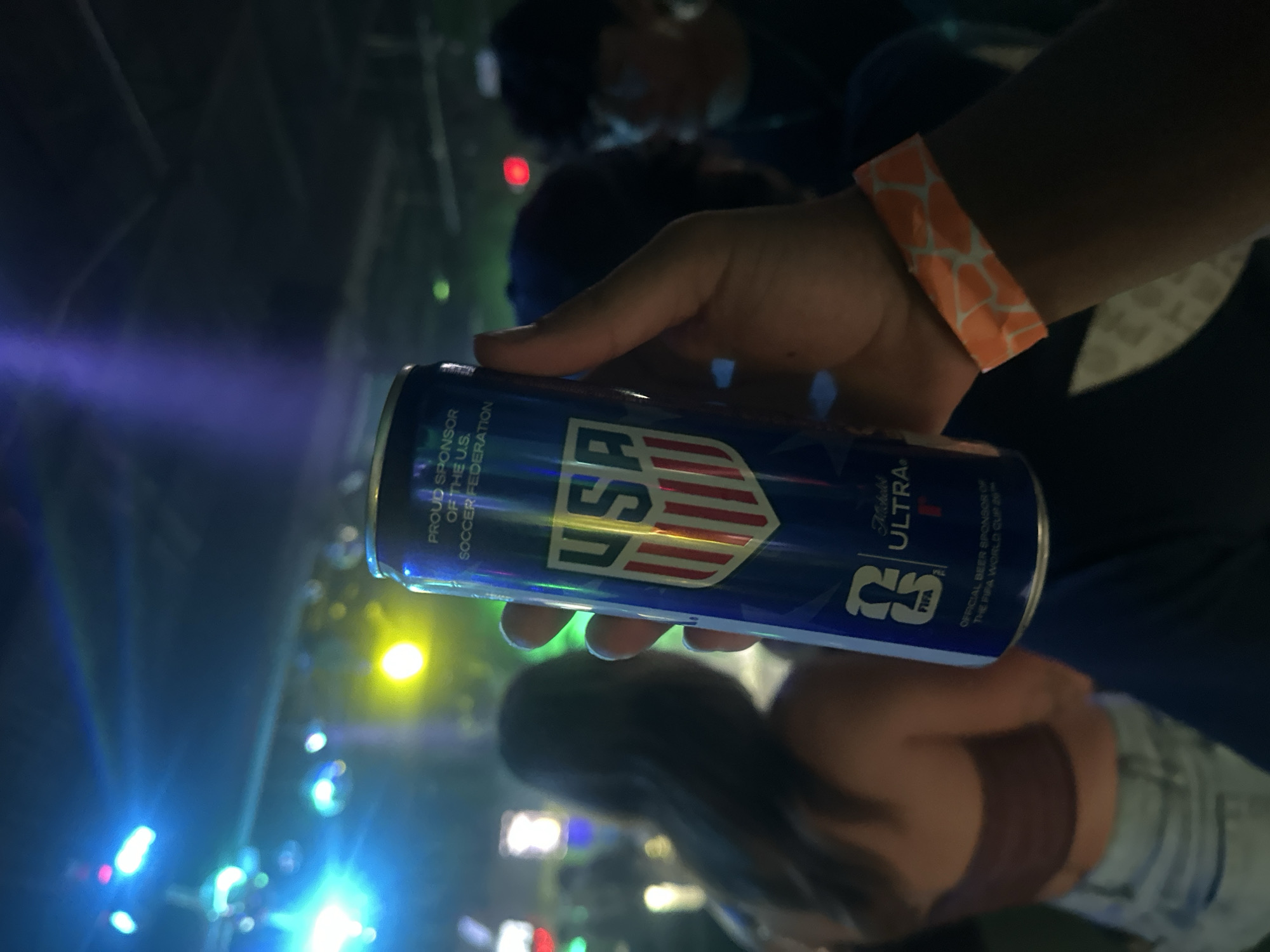 The media file presents a close-up shot of a person's hand holding a blue can at an indoor event, likely taking place at night in Tampa, United States. The can is prominently branded with the "USA" shield logo and identified as Michelob ULTRA. Visible text on the can indicates it is the "PROUD SPONSOR OF THE U.S. SOCCER FEDERATION" and the "OFFICIAL BEER SPONSOR OF THE FIFA WORLD CUP 26™". An orange and white patterned wristband is worn on the wrist holding the can, suggesting entry to a specific event or venue.
The background is dark and intentionally blurred, with numerous bright, colorful lights in shades of blue, yellow, red, and green creating a vibrant, energetic atmosphere. Silhouetted and blurred figures of other attendees are visible in the background, including a person wearing a bra-top and shorts, and another with dark hair. The overall scene suggests a lively social gathering, possibly a sports viewing party, concert, or celebration related to soccer, given the sponsorship details on the can.