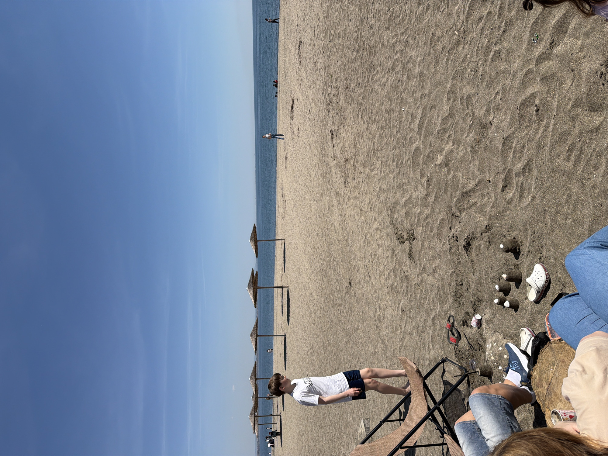 The image captures a beach scene in Unknown, Unknown. Predominantly, the environment consists of sand, a body of water, and clear sky. Several sun umbrellas are stationed along the beach near the water. In the mid-ground, a person is standing near a beach chair. Adjacent to the chair, there are several small objects on the sand, including a pair of white Crocs, blue New Balance sneakers, and a few small, white, bottle-shaped objects. The photograph also shows people in the water in the background.
