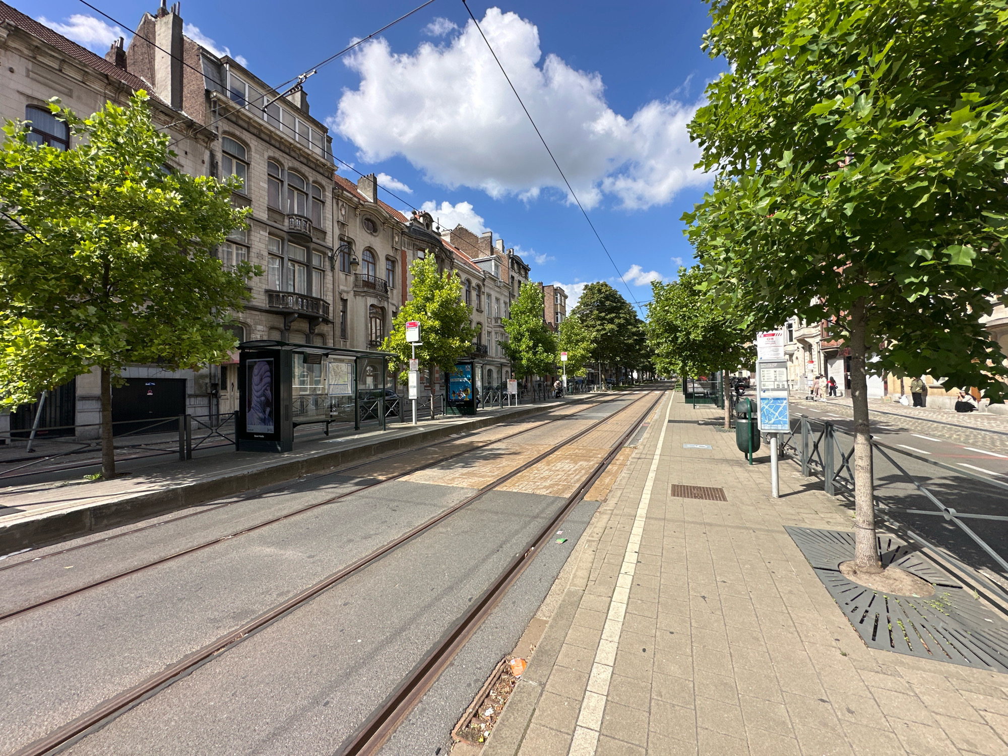 The image shows a street scene in Molenbeek-Saint-Jean - Sint-Jans-Molenbeek, Belgium, with tram tracks running down the center of the road. On the left, there are multi-story buildings with traditional architecture. A tree with green foliage is in the foreground on the left. On the right side of the street, there is a sidewalk lined with trees and lampposts. Several people are visible walking on the sidewalk and near a tram stop. The sky is blue with scattered white clouds. The overall impression is of a well-maintained urban street on a sunny day.