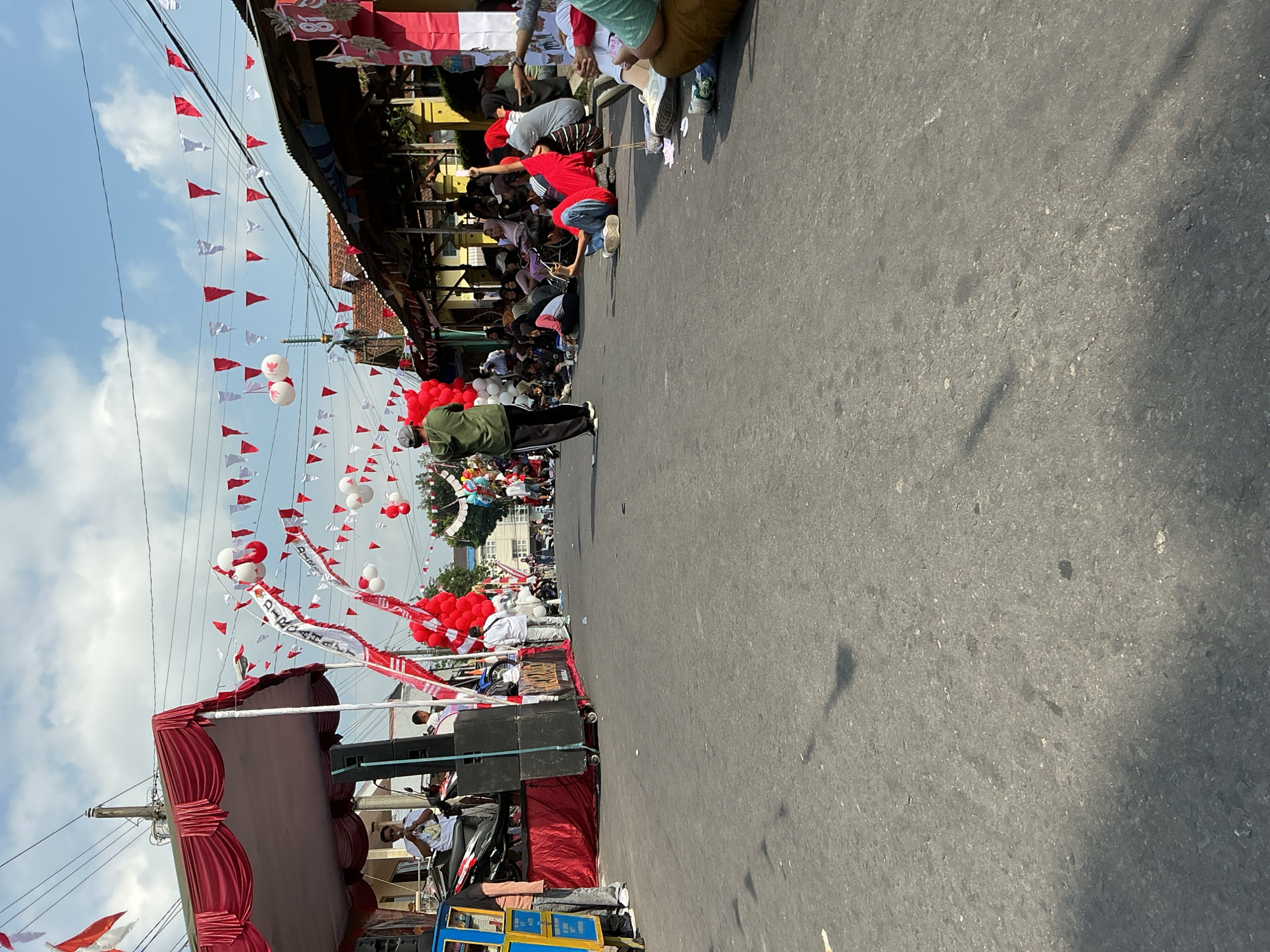 In the city of Magelang, Indonesia, a street scene unfolds on a bright, sunny day, dominated by red and white decorations, indicative of a national celebration. A street is cordoned off, and crowds of people are gathered on both sides, some seated on the asphalt. The atmosphere suggests a parade or a public event.
On the left side of the street, a red and white draped stage is set up, featuring a sound system with large speakers. A person is on stage, holding a microphone and appearing to address the crowd. Nearby, a colorful cart with a blue and yellow canopy stands. A number of red balloons are clustered at the base of the stage.
Across the street, a row of buildings with covered porches lines the road, with more people seated and standing in front of them. Festively strung red and white triangular flags and balloons crisscross the street overhead.
A person in a green hoodie stands in the middle of the street, facing away from the camera, holding what appears to be a small object. Other individuals are scattered throughout the street and sidewalk areas. The ground is asphalt, showing signs of wear and scattered debris. Shadows cast by overhead objects and buildings stretch across the road. The sky above is a bright blue with scattered white clouds.