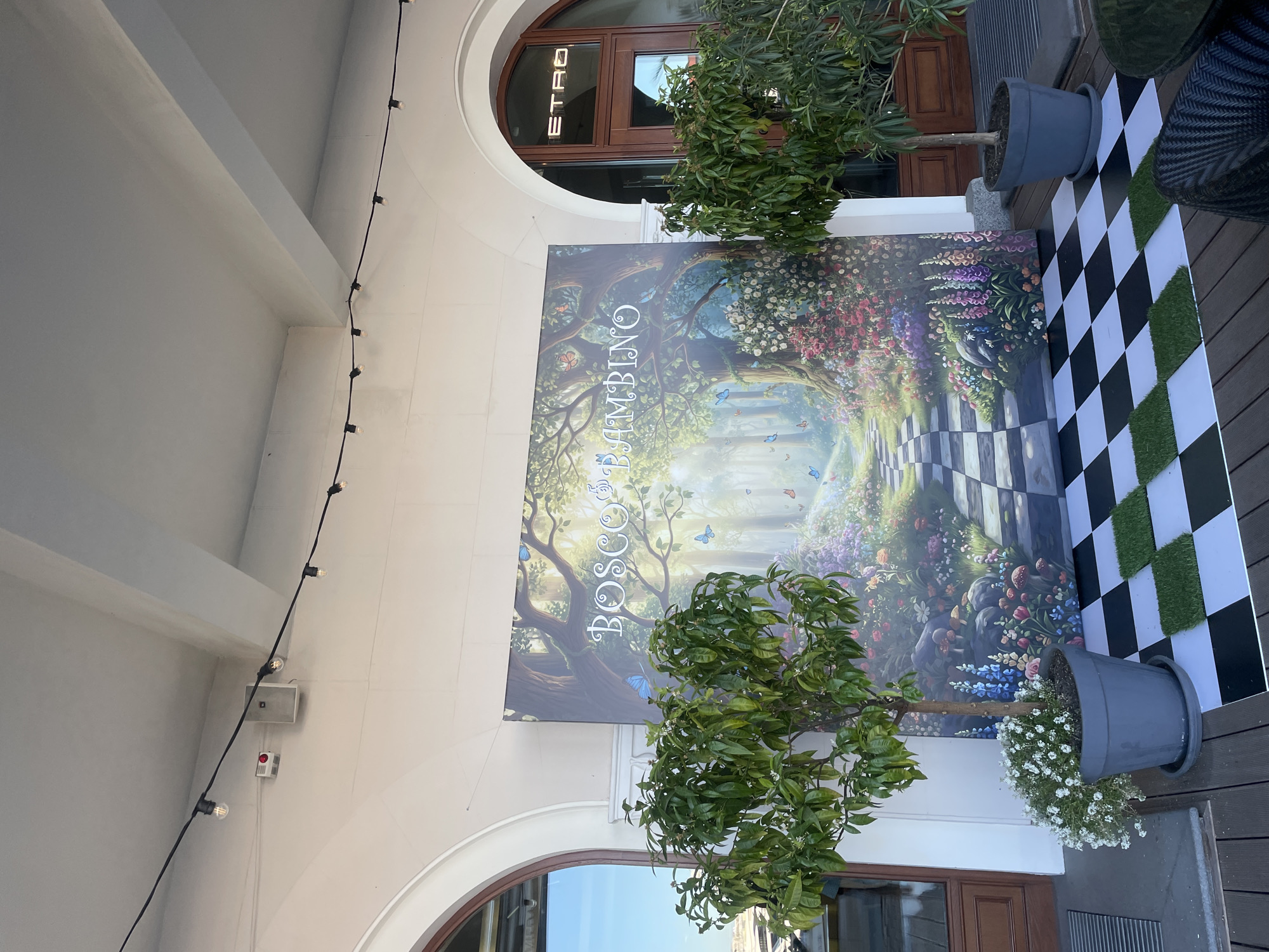 This media file depicts an ornate outdoor or semi-covered entrance area, likely to a shop or establishment. A large, vibrant mural dominates the scene, illustrating a fantastical, lush garden with rich greenery, diverse colorful flowers, and numerous blue butterflies fluttering amidst the foliage. A winding path, made of checkered stones, leads into the whimsical forest depicted on the mural.
Prominently displayed on the mural is the text "BOSCO BAMBINO" in an elegant, stylized font. In front of the mural, on a wooden deck, a decorative black and white checkered mat or flooring is laid out, interspersed with squares of artificial green grass, creating a striking visual contrast. Two potted small trees, one with delicate white flowers, are placed on either side of the mural, contributing to the natural aesthetic.
The architectural setting features a white, arched facade. Through an arched window or doorway in the background, the illuminated text "ETRO" is visible, indicating a luxury retail brand. A string of small, warm-toned light bulbs hangs overhead, suggesting decorative lighting. Additional details include a grey speaker and a red button mounted on the white wall. The scene is brightly lit, indicating daytime. No people are visible, and no specific activity is taking place. The setting is in Sochi, Russia.