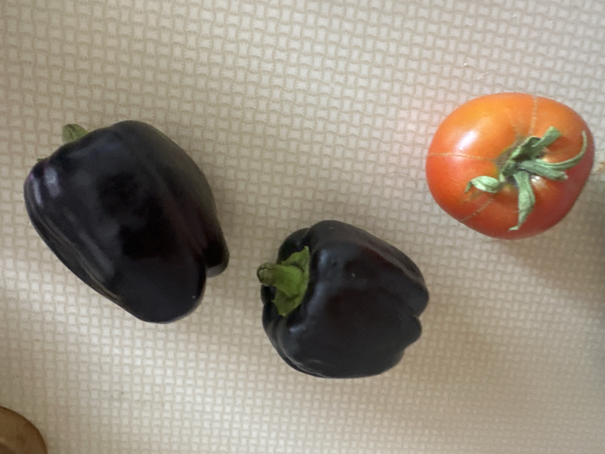 Three bell peppers, one red tomato, and a wooden object are on a light-colored, textured surface. The bell peppers are dark purple and have green stems. The tomato is red with green stems and appears to be slightly bruised. The wooden object is partially visible in the upper right corner of the image. The scene is indoors, likely on a table or countertop. The location is not specified in the image.