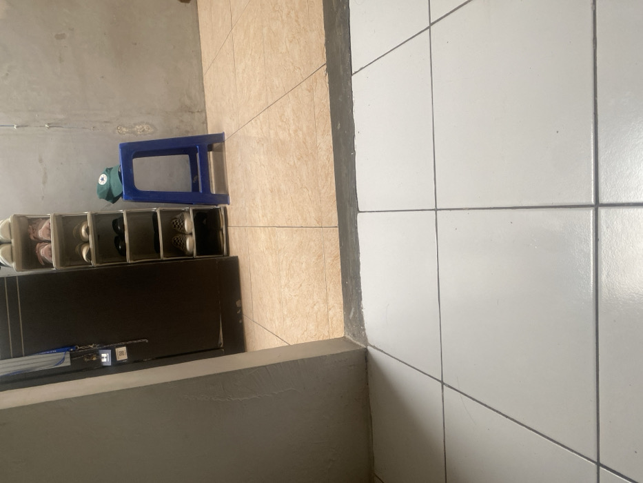 The image depicts an interior scene, observed from a low vantage point across a transition between two distinct floor areas, located in Cimenyan, Indonesia. In the foreground, the floor consists of large square white ceramic tiles with dark grey grout lines. A dark grey or black concrete threshold separates this area from the room beyond.
The room beyond the threshold features light brown/beige ceramic floor tiles. To the left, a dark brown, vertically oriented structure, possibly a cabinet or doorframe, is partially visible. Attached to its left side are a blue lanyard and a small white rectangular object displaying what appears to be a QR code or barcode. Adjacent to the right of this structure stands a narrow, tall shoe rack with six open shelves. From top to bottom, the shelves hold: two pairs of light-colored athletic shoes, two pairs of pink athletic shoes, two pairs of light brown/beige shoes, two pairs of black closed-toe shoes, two pairs of checkered pattern slip-on shoes, and a single dark shoe on the bottom shelf.
To the right of the shoe rack, a solid blue plastic four-legged stool is positioned against a plain, light grey concrete or stucco wall. A green baseball cap with a white star logo is resting on the seat of the blue stool. The overall environment appears well-lit by ambient light. No individuals or active interactions are present in the scene.