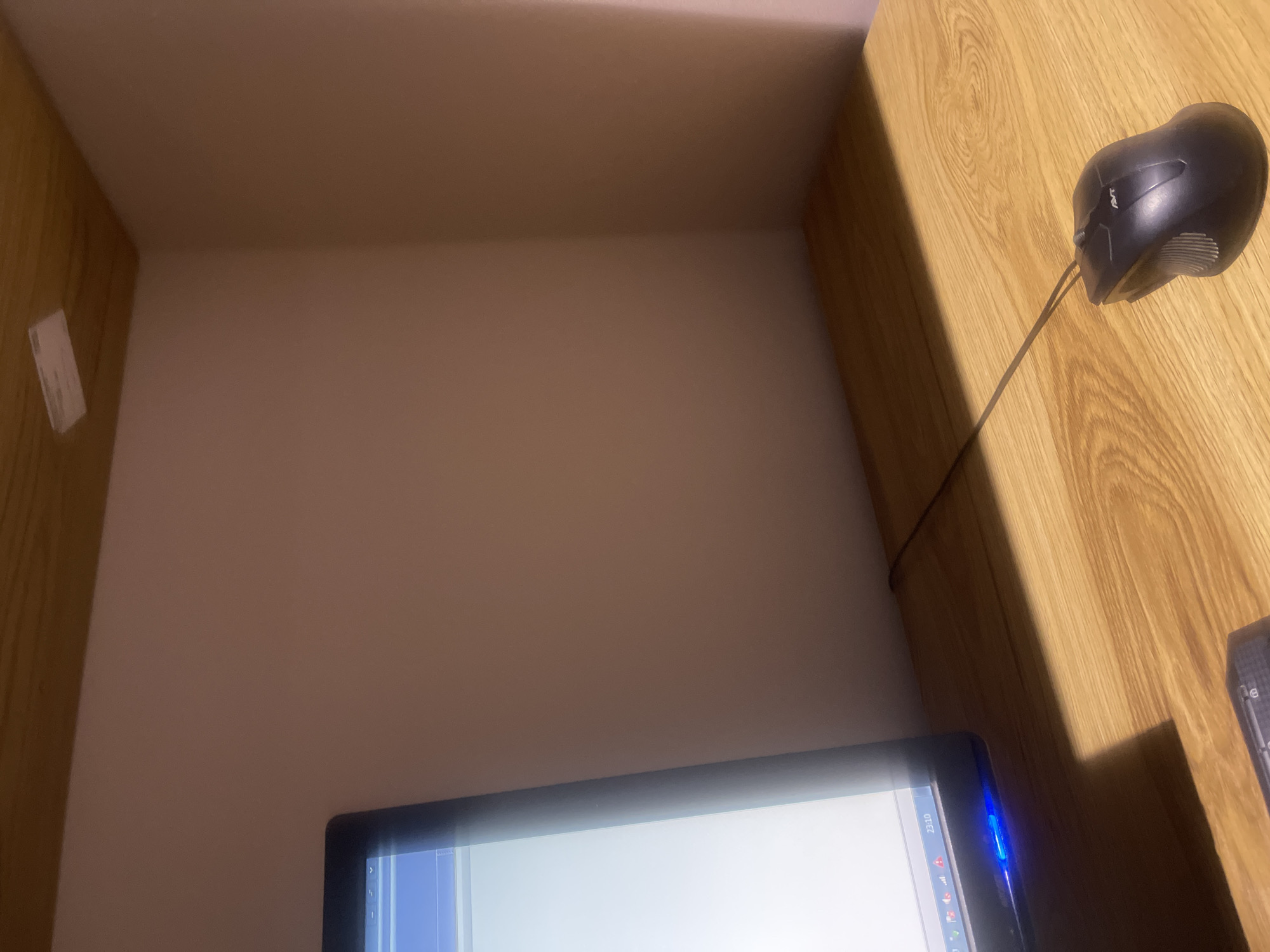 A computer mouse rests on a wooden desk in front of a computer monitor. The mouse is black and has a wire connecting it to the computer. The desk has a wood grain pattern. The monitor is on the left side of the frame and displays a white screen with some icons. The background is a plain wall. The scene appears to be indoors, possibly in an office or a home study. The lighting is dim, suggesting it might be nighttime or the room is not well-lit. There is no indication of the city of Qarshi, Uzbekistan, or any specific location.