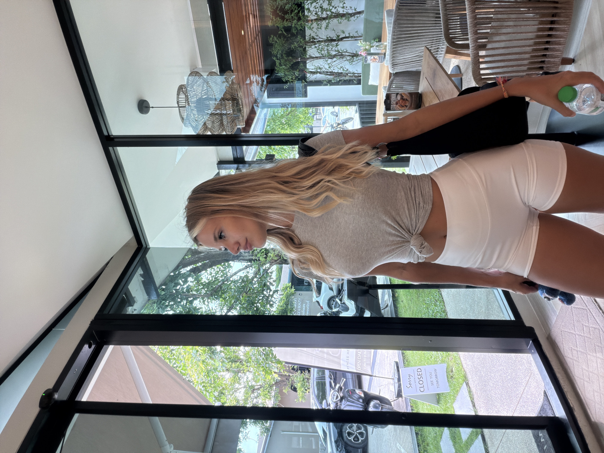 A blonde woman is standing in front of large glass doors, looking to her right. She is wearing a grey t-shirt tied at the waist and white shorts. A black bag is slung over her right shoulder. In her right hand, she is holding a water bottle. A small blue toy is in her left hand. The doors appear to lead to an indoor restaurant, with tables and chairs visible inside. Outside the doors, there is a paved area with grass and trees. A black scooter and cars are parked on the left side, and a sign that reads "SORRY CLOSED SEE YOU TOMORROW" is visible on the glass door. The lighting suggests it is daytime. The location is identified as Si Sunthon, Thailand.