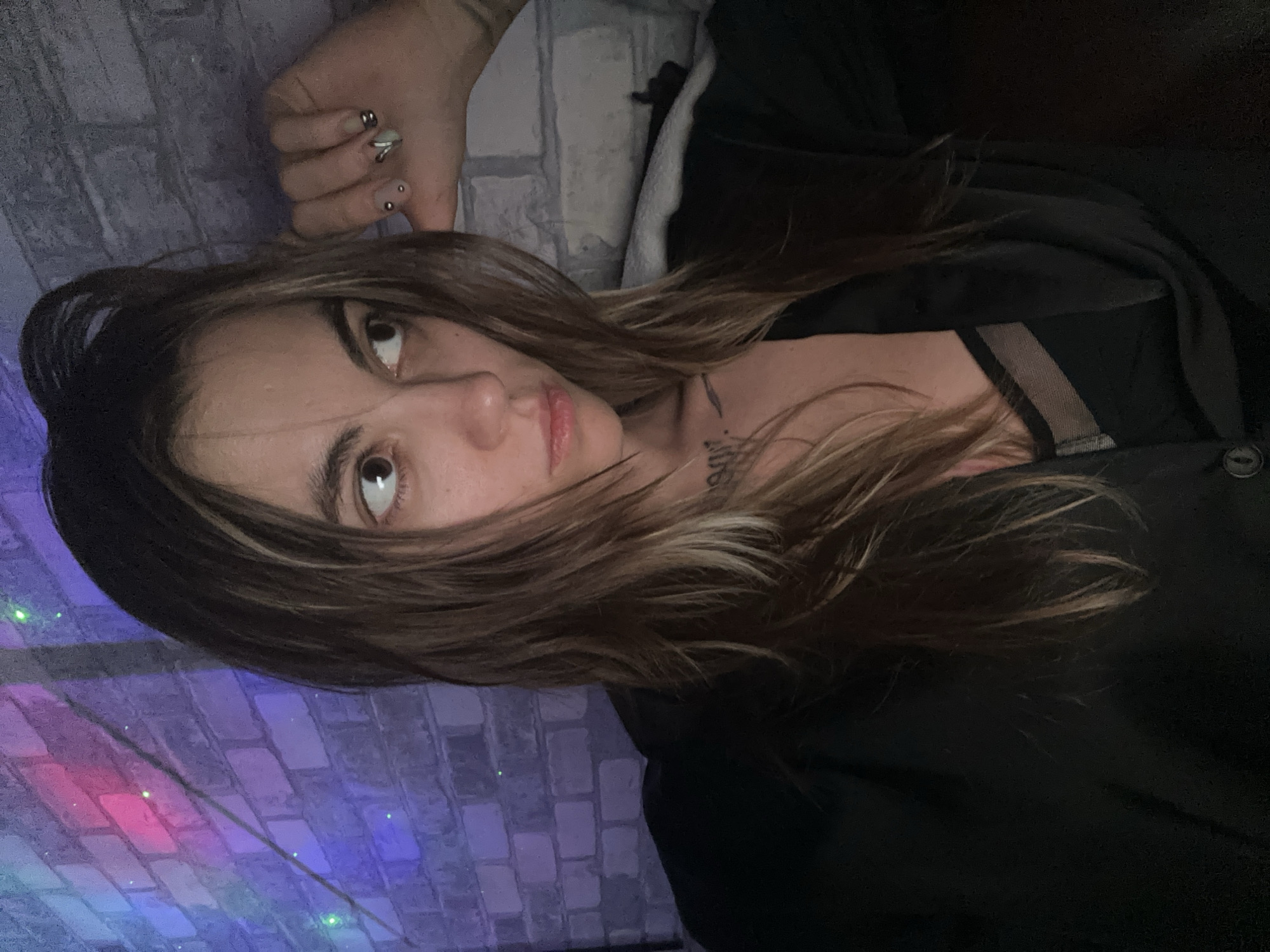 A young woman with long, light brown hair, pulled back on top, is captured in a medium-close up selfie. She is looking to her right with a neutral expression. She is wearing a black garment, possibly a jacket or blouse, over a black lace bra. She has a tattoo on her chest and her fingernails are painted with a light green polish and adorned with small silver embellishments. Her right hand is raised and resting on her head, revealing a silver ring on her index finger. The background is a brick wall illuminated with colorful lights, creating a vibrant and somewhat moody atmosphere with hues of pink, purple, and green. The image appears to be taken indoors, in what could be a home or a dimly lit venue, likely at night. No specific location in Moscow, Russia can be determined from the visual information provided.