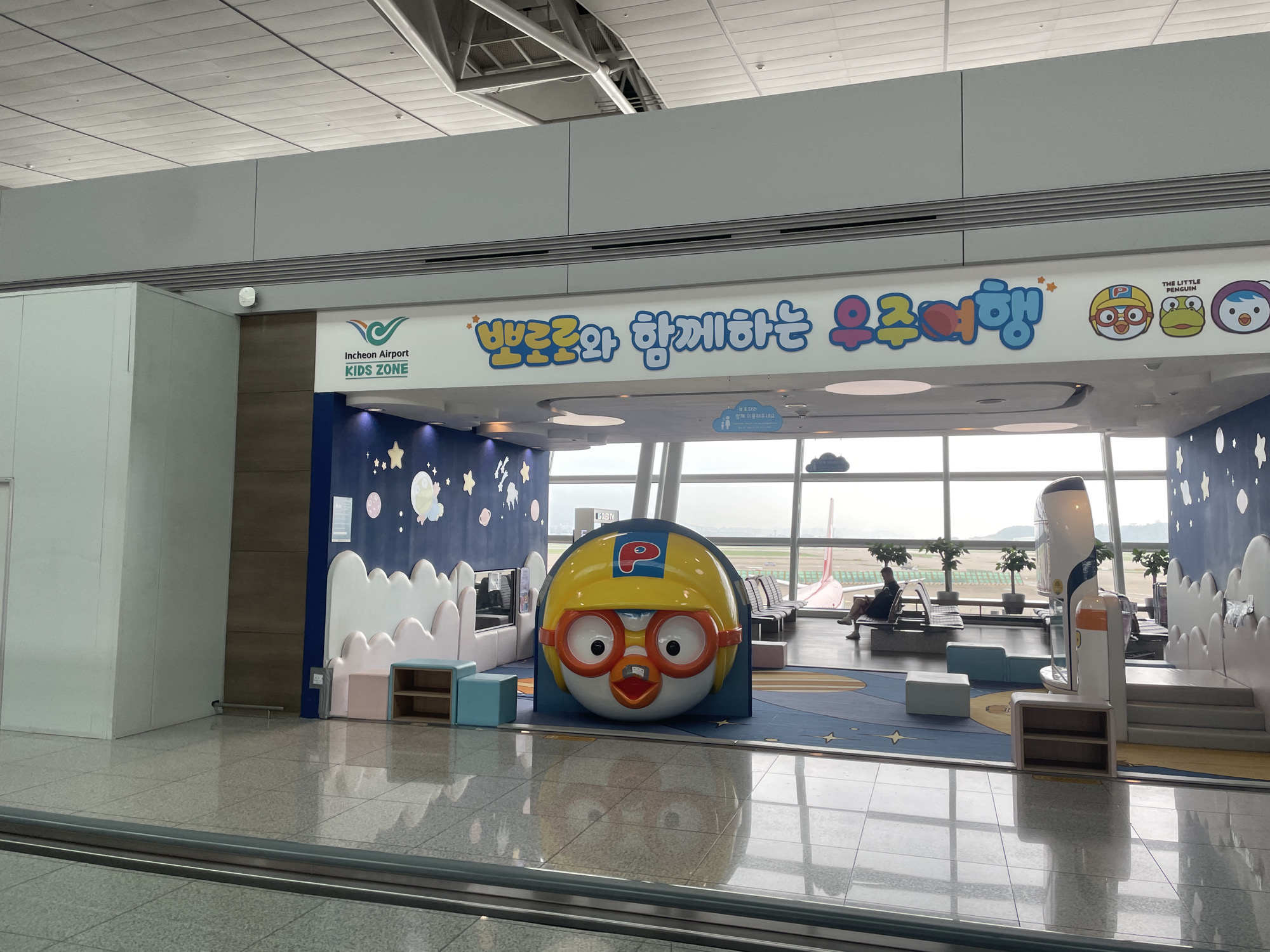 This is an indoor photograph of the Incheon Airport Kids Zone, located in Incheon, South Korea. The image captures a brightly decorated play area designed for children. A large, cartoonish character head, identified as Pororo the Little Penguin, is prominently displayed at the center of the scene. The character is wearing a yellow helmet and goggles, with an open mouth and large eyes. Behind Pororo, a wall is adorned with a blue night sky mural featuring white clouds, stars, and planets.
To the left of the main character, a sign reads "Incheon Airport KIDS ZONE" with a stylized green leaf logo above it. The play area itself is carpeted with a blue mat featuring a colorful space-themed design, including stars and planets. Several low, white cloud-shaped structures are present, serving as decorative elements and possibly seating or play surfaces. Small, light blue and pink cube-shaped seats are also visible.
In the background, large windows offer a view of the airport tarmac. Several rows of airport seating can be seen through the windows, and a person is sitting on one of the benches. A part of an airplane wing is visible outside the window, indicating its proximity to the airport operations. The floor in the foreground is polished and reflective, showing the reflection of the play area and the overhead lighting. The ceiling is a typical airport ceiling with acoustic tiles and exposed structural elements. The overall atmosphere suggests a family-friendly space within the airport.