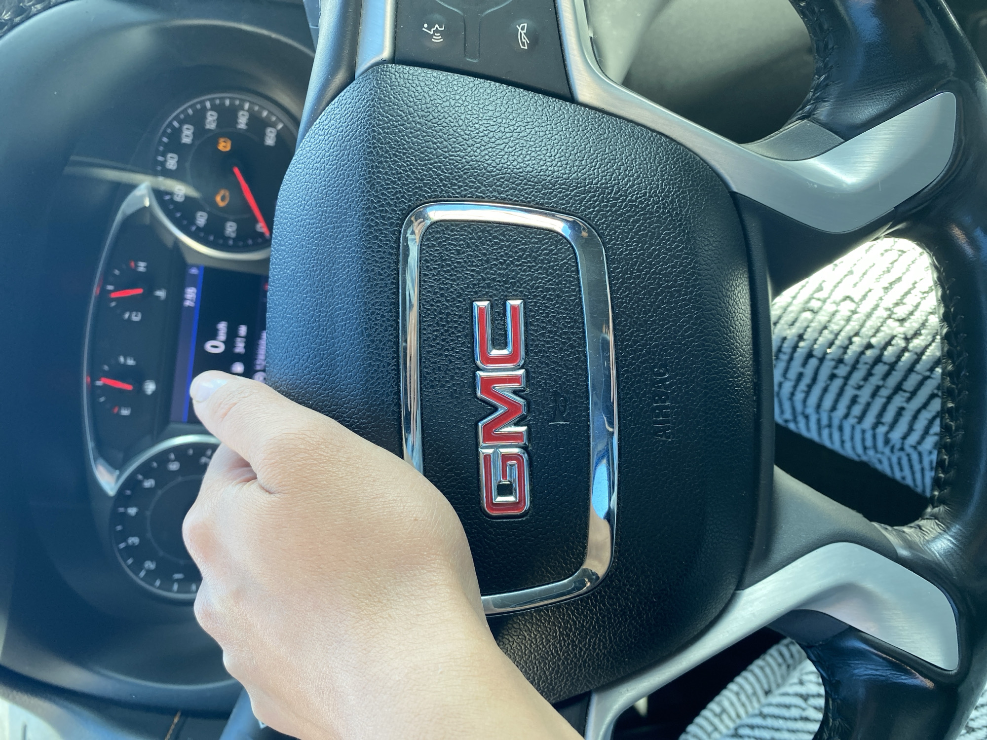 A person's hand rests on the steering wheel of a GMC vehicle. The steering wheel has a textured black grip and a silver trim surrounding the GMC logo. On the dashboard, a digital display shows "0 km/h" and "7:56," indicating the vehicle is stopped or moving at a very low speed. There are also warning lights illuminated on the dashboard, including a check engine light and a battery light. The background shows the car's interior. The location is Unknown, Unknown.