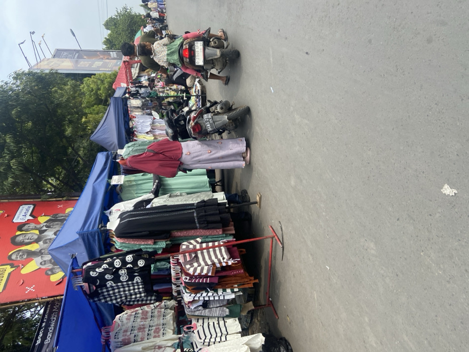 The image shows a street market in Margaasih, Indonesia (latitude: -6.953, longitude: 107.564). Several vendors have set up shop along the side of the street, displaying clothing items on racks. There are several customers browsing through the wares. One woman is standing in front of a clothing rack, wearing a long, pink hijab, examining the items. A man and a young girl are standing on the side of the street, looking at the merchandise. The man is wearing a red shirt and a backpack. The girl is wearing a pink dress. Behind them is a motorbike. The street is paved with asphalt. The sun is shining and the sky is cloudy. There are trees and buildings in the background.