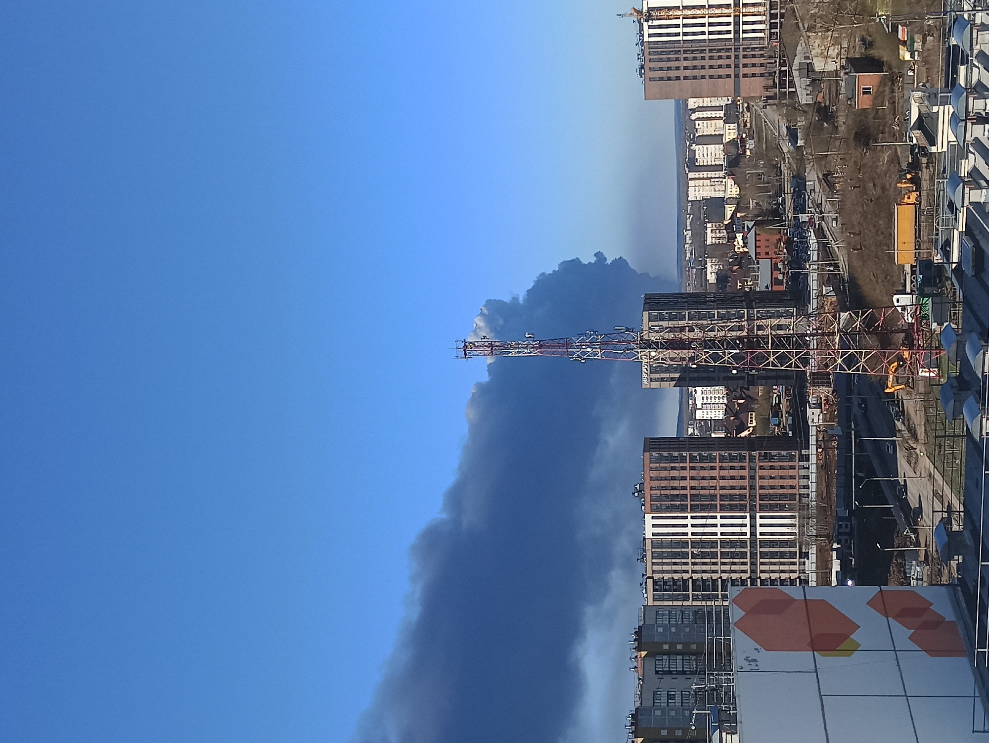A medium-angle daytime shot depicts a city scene, with a focus on dark smoke rising into a clear blue sky. The location is Unknown, Unknown.
In the foreground, urban buildings are visible, some of which feature white or light-colored ventilation systems. A tall, lattice-like metal structure rises amid the buildings, contrasting with the smoke column.
The smoke plume is significant, indicating a fire or other event producing substantial emissions. The background features additional city buildings, roads, and some green areas. Construction equipment is visible, including a yellow crane and a yellow vehicle on the right side of the road.