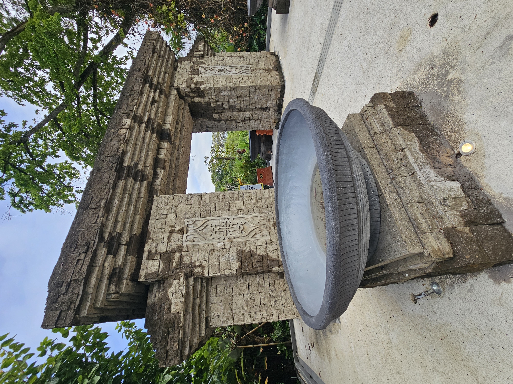 The image depicts a stone gateway structure in Denpasar, Indonesia. The structure is made of light-colored stones and features decorative carvings. A large, round planter is situated to the side of the gateway. Through the gateway, one can see greenery and buildings. The scene appears to be outdoors, possibly in a garden or park setting. The presence of a small light fixture indicates the area may be illuminated at night. The image is oriented with the top of the scene tilted to the left.