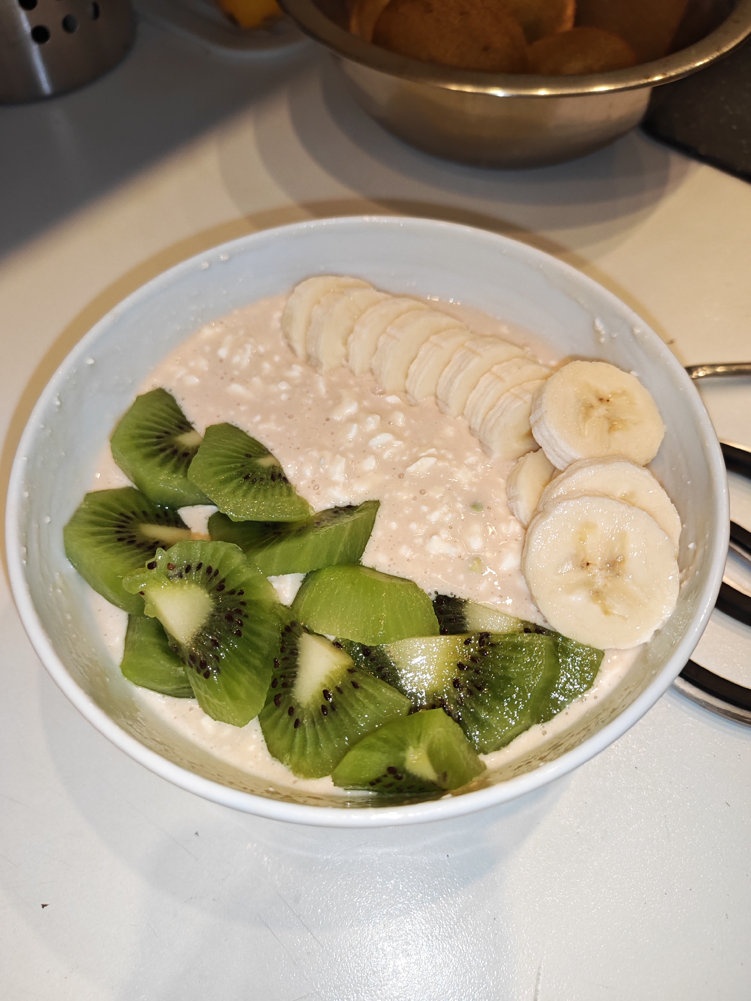 A white bowl contains a creamy, light pink mixture topped with sliced bananas and kiwi. The kiwi slices are arranged on the left side of the bowl, with their green skins and black seeds visible. The banana slices are on the right side, forming a curved line. The bowl is on a white surface, likely a countertop, with a metallic container and other food items blurred in the background. The lighting suggests an indoor setting. The image does not contain information about the city of Bataysk, Russia.