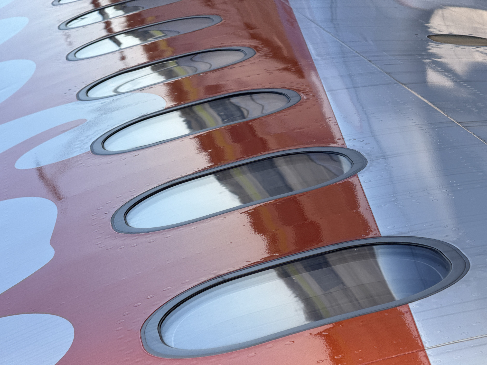 The image shows a portion of an aircraft's wing featuring a row of oval-shaped windows. The wing is painted with a combination of red-orange and white. The exterior of the wing appears glossy, potentially due to a wet surface or a reflective coating. The windows reflect the surrounding environment. The airframe construction exhibits rivets visible on the metallic portion of the wing. The location in the city of Niederanven, Luxembourg is not evident from the image.