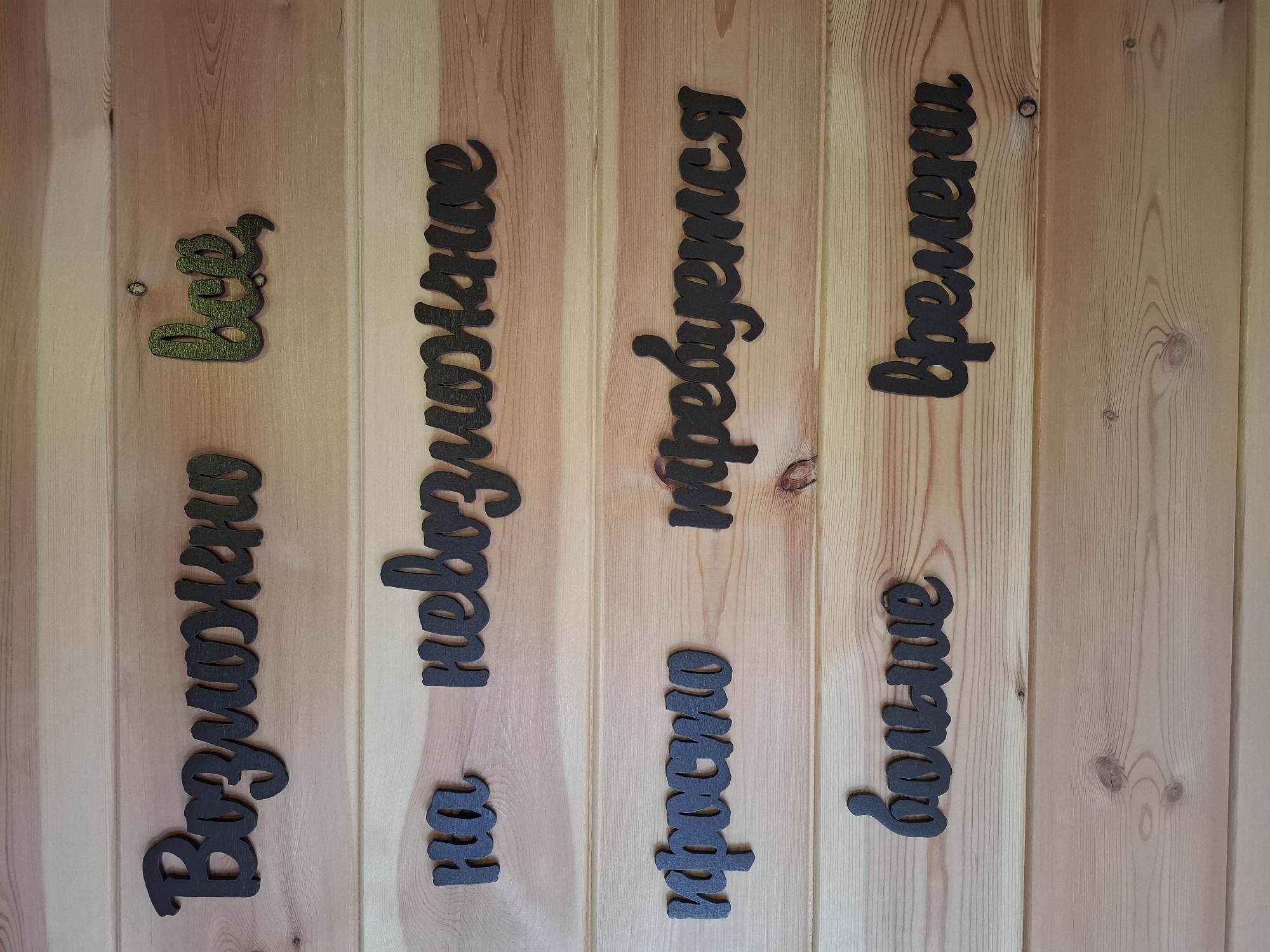 The image shows a wooden wall with several phrases written in Russian, made of black, cut-out letters, possibly wood or plastic. The phrases are arranged on separate wooden planks.
From top to bottom, left to right, the phrases are:
- "Возможно" (Vozmozhno - Possibly)
- "все" (vse - everything) - this word is green with a textured surface.
- "на" (na - on)
- "невозможное" (nevozmozhnoye - impossible)
- "просто" (prosto - simply)
- "требуется" (trebuyetsya - requires)
- "больше" (bol'she - more)
- "времени" (vremeni - time)
The wall appears to be made of light-colored wooden planks, possibly pine, with visible grain and knots. The lighting suggests an indoor environment, possibly a room or a covered outdoor area. The overall aesthetic is rustic and decorative.
There are no people or animals visible in the image. The scene is static and appears to be a close-up shot of the wall art. The context of Sukhoy Ruchey, Russia is not visually evident from the image itself.