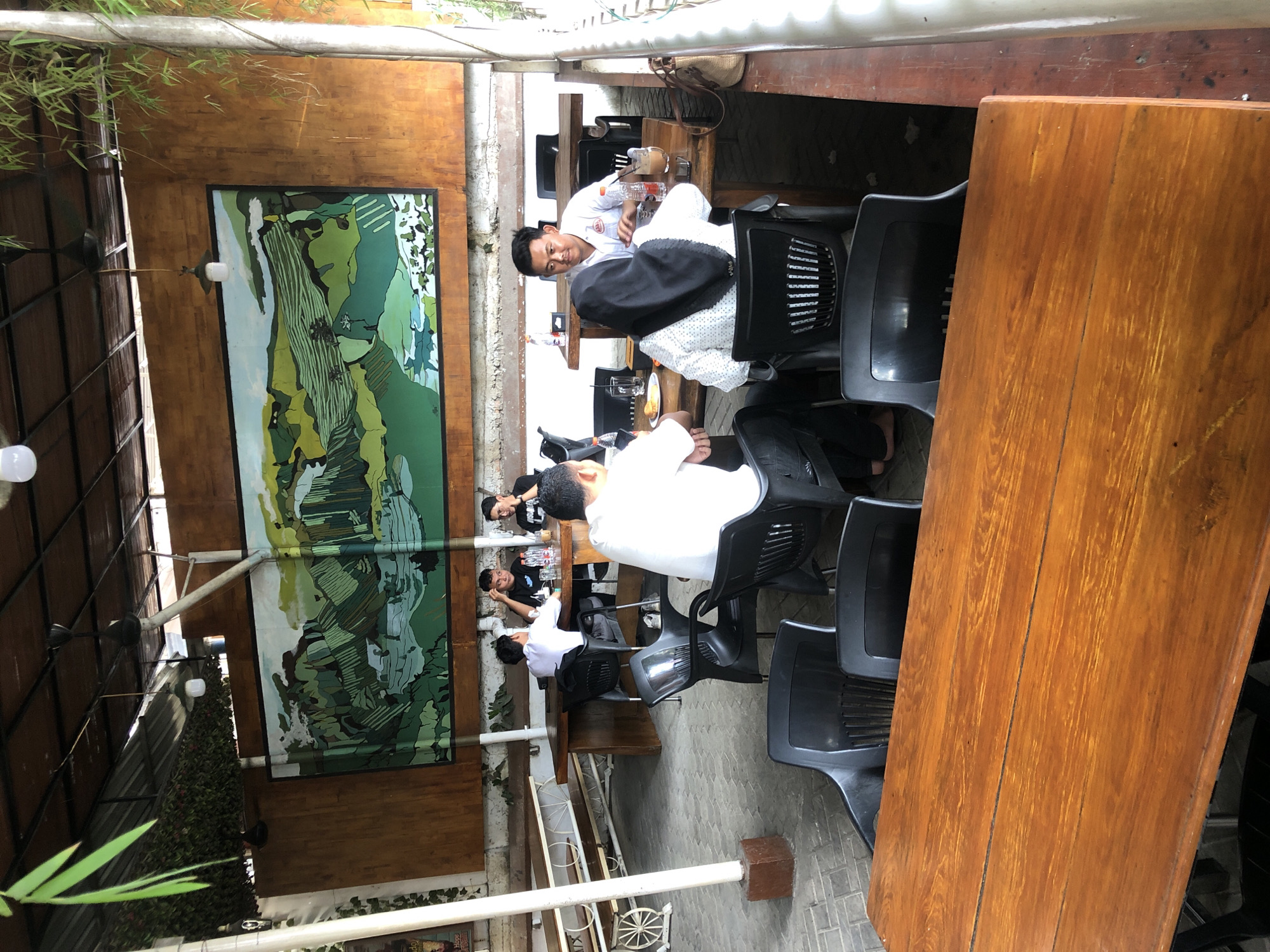 This image depicts an outdoor seating area at a cafe or restaurant. The setting appears to be the city of Langsa, Indonesia. In the foreground, a polished wooden table is prominent. Behind it, black plastic chairs are arranged.
Further back, a group of people are seated at tables, engaged in conversation and consuming drinks. One table has three men, two of whom are wearing black t-shirts and one in a white shirt, with water bottles and glasses on the table. Another table features two individuals: a man in a white shirt and a woman wearing a black hijab and a patterned white shirt. They have glasses, a bottle of water, and a coffee drink between them.
The environment is an open-air space with a covered roof supported by beams and poles. A large, vibrant mural of a green landscape, possibly depicting terraced fields, adorns the wall behind the seating area. Some greenery, including hanging plants and bamboo, is visible, adding to the outdoor ambiance. The floor is paved with light-colored bricks. The overall scene suggests a casual dining or social gathering.
