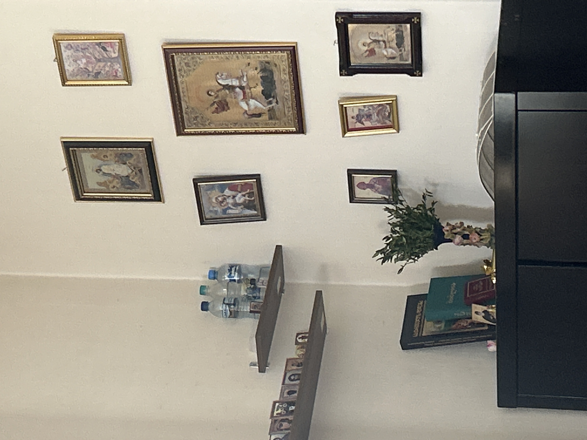 In the city of Unknown, Unknown, an interior room corner is depicted, featuring light-colored walls. On the left wall, two wooden floating shelves are mounted. The upper shelf holds three plastic water bottles with blue and green caps on its right side, alongside a small framed icon. The lower shelf displays a linear arrangement of multiple small framed pictures or icons. The right wall is adorned with seven framed pictures or paintings, primarily religious icons or classical art, arranged in three rows. The largest central painting depicts a figure on a white horse, set against a golden background. In the foreground, a dark-colored cabinet or dresser presents a flat top surface. On this surface, a dark-bound book stands upright, adjacent to a teal-colored hardcover book. A small framed icon of a person is positioned in front of a reddish-brown book. To the right, a dark vase containing green foliage and decorated with floral patterns is present. A subtle, curved, ribbed object is partially visible on the far right of the furniture's surface. No individuals are present, and no actions are observed.