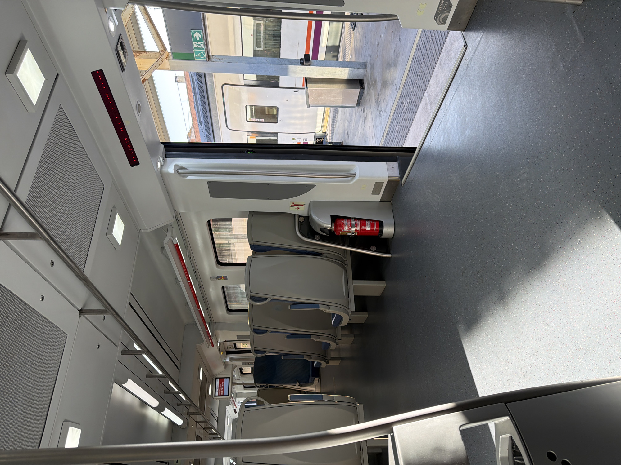 The image shows the interior of an empty train carriage, with a view through an open doorway to a train on an adjacent platform. The train carriage is modern and clean, with grey seats arranged in rows and a grey floor. There is a fire extinguisher mounted on the wall near the open doorway. Outside the train, a station platform is visible, with a silver trash bin and a sign indicating accessibility and an exit. The train on the platform is white with a purple and orange stripe. The lighting suggests it is daytime, with sunlight entering the carriage and casting shadows on the floor. Based on the red sign displayed inside the carriage that reads "DESTINACIÓ: PORTBOU," the location is likely a train station in or near Figueres, Spain, as Portbou is a town on the French border accessible by train from Figueres.