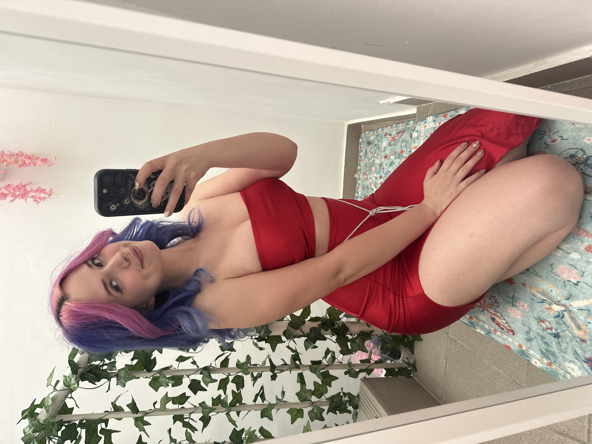 An indoor mirror selfie captures a young woman with striking multi-toned hair, featuring shades of pink and blue/purple. She wears a form-fitting, strapless red two-piece outfit, consisting of a top and what appears to be a matching skirt or high-waisted bottoms, accentuated by a decorative silver or white beaded string detail on her side. Her nails are manicured with a light, reflective polish.
The woman is smiling subtly as she looks into the mirror, holding a smartphone with a dark, intricately designed case – possibly featuring a large eye motif – in her left hand to take the picture. Her right hand rests gently on her right thigh. She is seated or kneeling on a patterned rug, which features colorful designs of butterflies and flowers in hues of blue, green, and red.
The setting appears to be a brightly lit room during the daytime. The background, seen in the mirror, includes a plain white wall with a small arrangement of pink artificial flowers on a shelf to the left. To her right, an artificial ivy garland is draped over a white frame or structure, adding a natural element. Adjacent to the rug, a light-colored tiled floor is visible. The image conveys a casual and personal moment. No specific text is visible in the frame, and while the location context is provided as Zagreb, Croatia, there are no explicit visual cues within the image to confirm this.