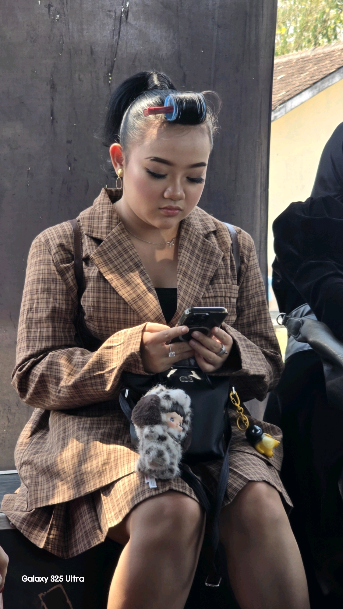 A young Indonesian woman with dark hair pulled into a ponytail and hair curlers on her front hair is seated outdoors, looking down at her smartphone. She is wearing a brown plaid blazer over a black top and a matching pleated skirt. A small black bag is slung across her body, adorned with a fuzzy charm and a yellow penguin keychain. The background consists of a dark, textured wall and a building with a yellow facade and a tiled roof. The image has a watermark in the lower left corner that reads "Galaxy S25 Ultra". The location is specified as Toroh, Indonesia.