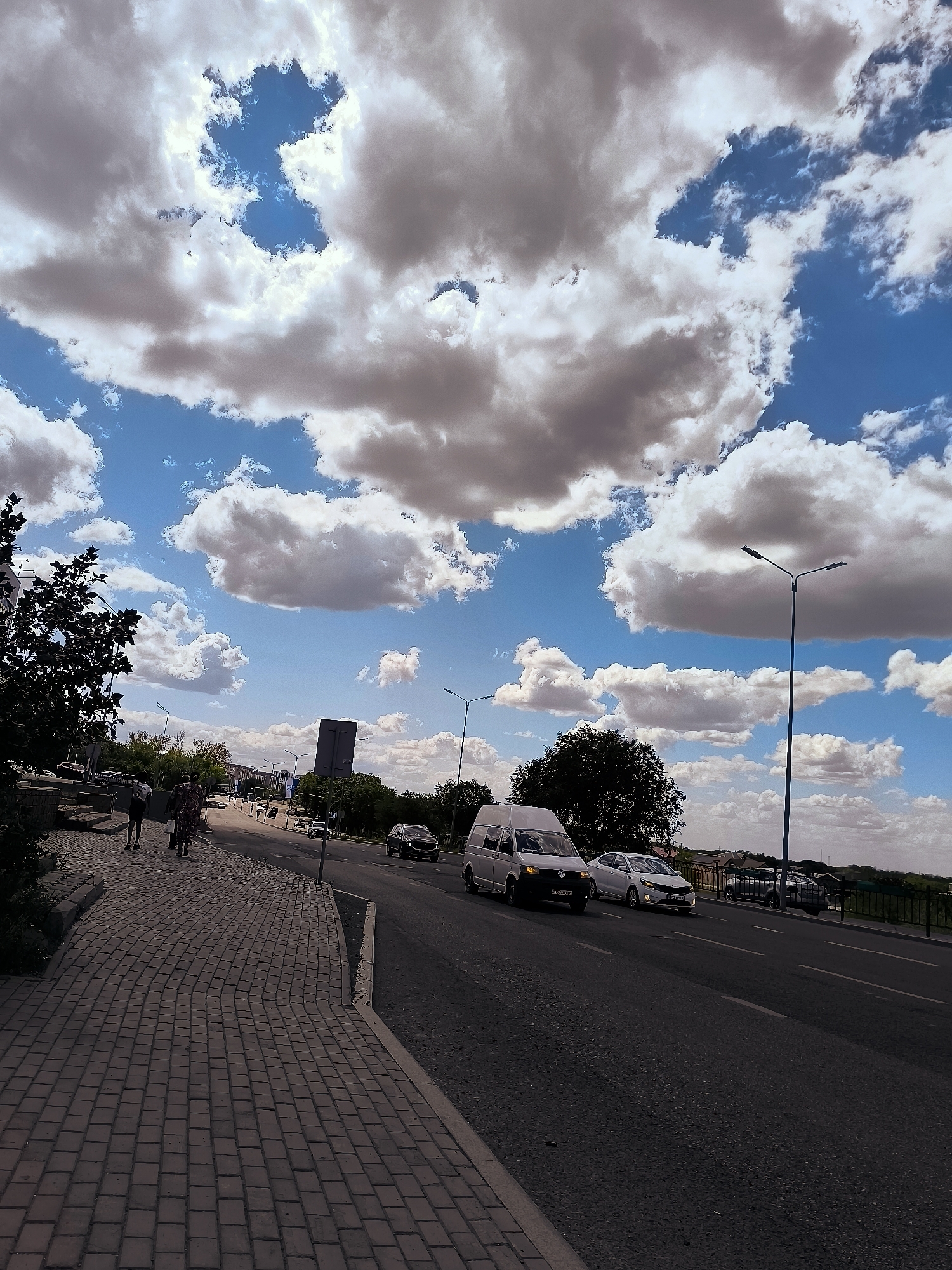 The image depicts a street scene in Qarağandı, Kazakhstan, with the city administration building visible in the distance. The sky is a vibrant blue, populated with large, fluffy white and grey clouds, suggesting a dynamic weather day. In the foreground, a paved sidewalk made of grey bricks runs along the left side of the image. Two individuals are walking on the sidewalk, facing away from the camera and towards the street. One person appears to be wearing darker clothing, while the other is in a brightly colored, patterned dress. They are carrying bags.
The street itself is asphalt and appears to be a multi-lane road with white lane markings. Several vehicles are present: a white van is driving in the right lane, followed by a white car. Further down the road, other vehicles are visible, including a dark SUV on the right side. Streetlights are positioned along the road, with one prominent lamp post on the right side of the image. Trees with dark green foliage line the sides of the road and sidewalk, contributing to the urban landscape. In the background, several buildings are visible, including a tall, dark structure that could be the city administration building, surrounded by other lower-rise structures and residential areas. The overall impression is of a typical urban environment under a dramatic sky.