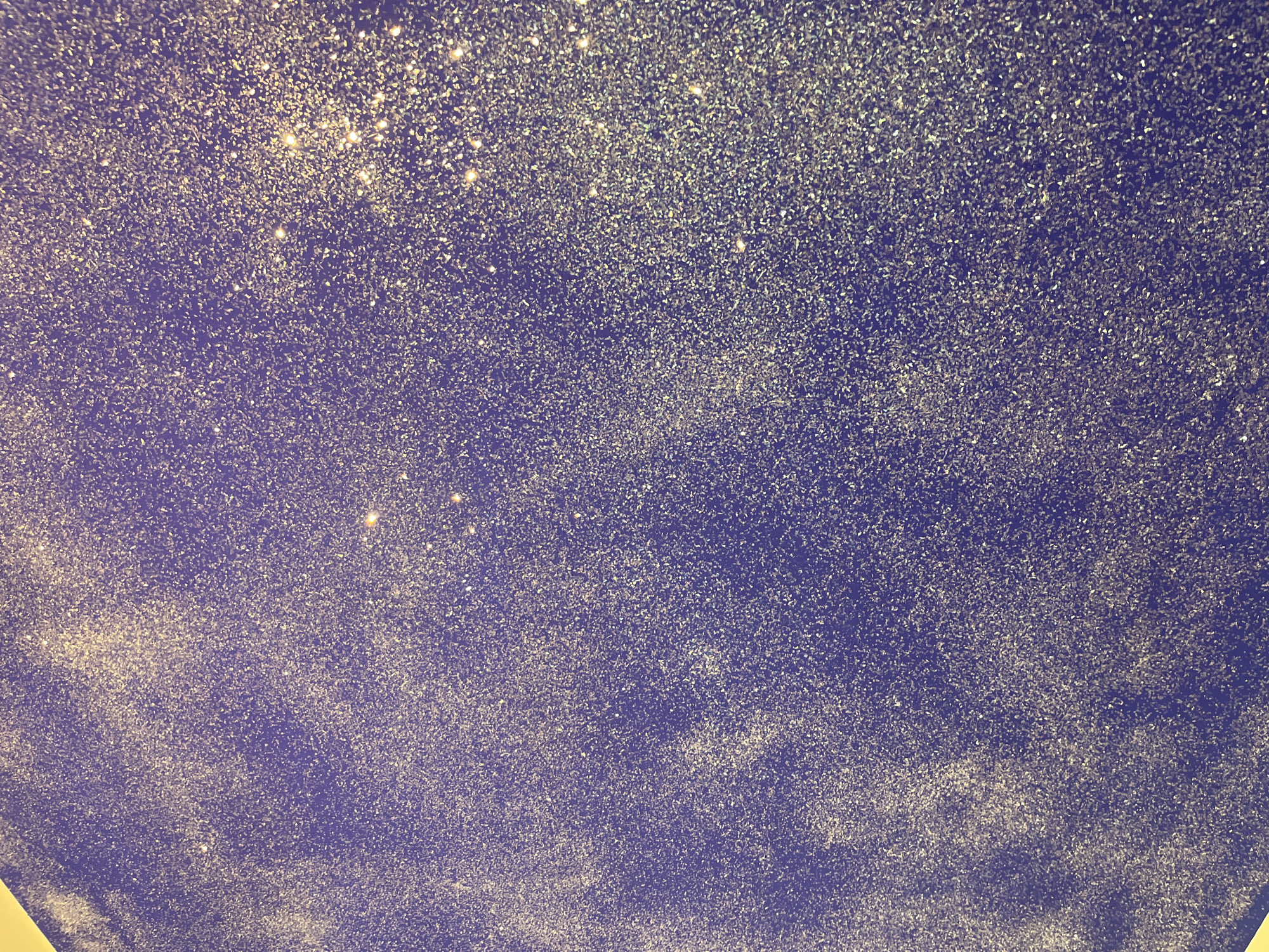 The image presents a textured surface in shades of blue and gold, resembling a night sky speckled with stars or a canvas with scattered glitter. The density of the golden specks varies, with heavier concentrations towards the top and bottom of the frame, suggesting a gradient effect. No people, specific objects, or identifiable actions are discernible in the image. The location is unknown.