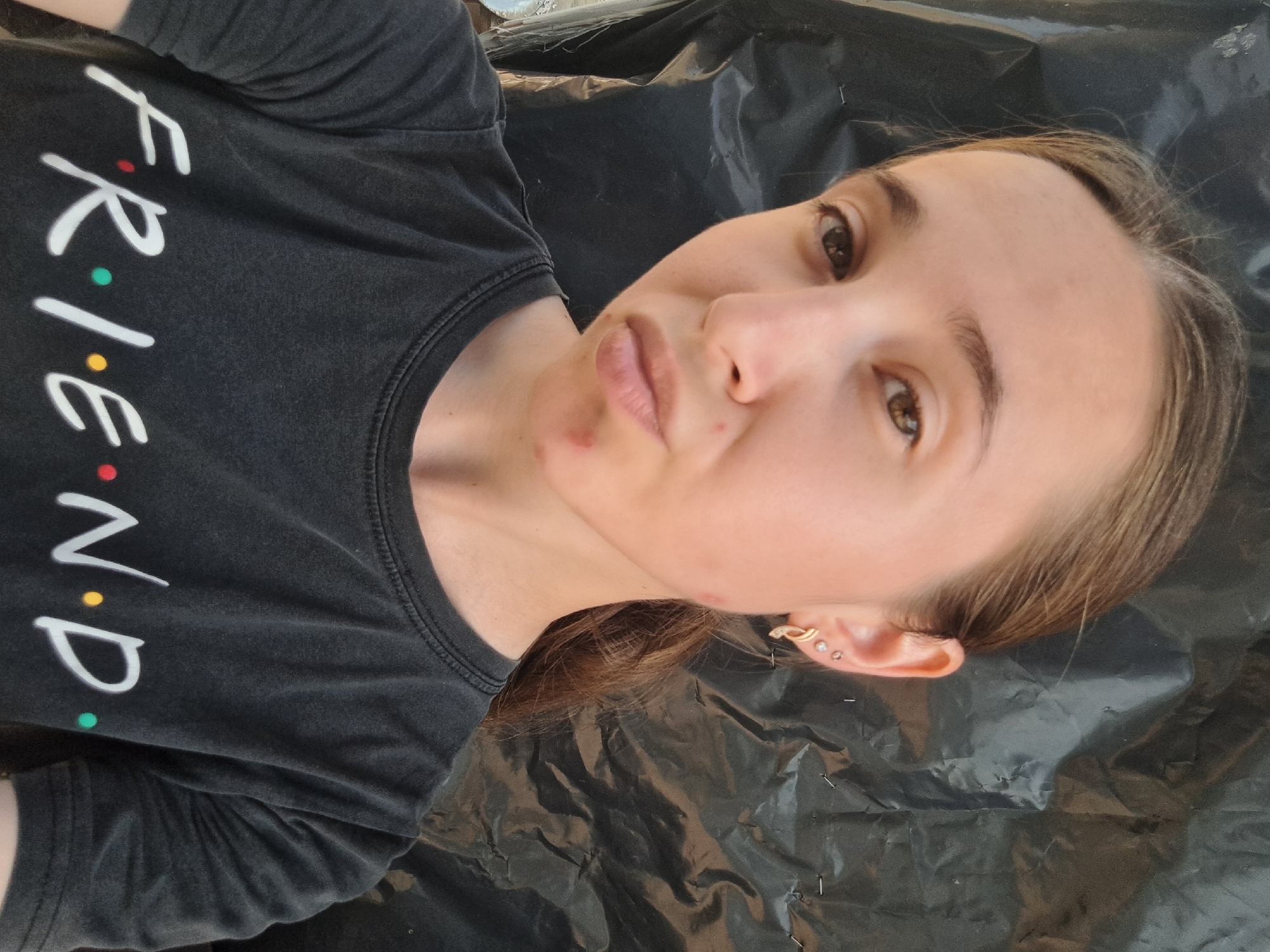 A young woman is taking a selfie in front of a black, crinkled backdrop. She has light brown hair pulled back into a ponytail and is wearing a dark gray t-shirt with the word "FRIENDS" printed on it in white letters, with colorful dots representing the vowels. She has two earrings in her left ear: a small stud and a hoop earring. There are a few small red blemishes on her chin. The backdrop appears to be some sort of plastic sheeting. The setting is not identifiable as being in the city of Vypasne, Ukraine.