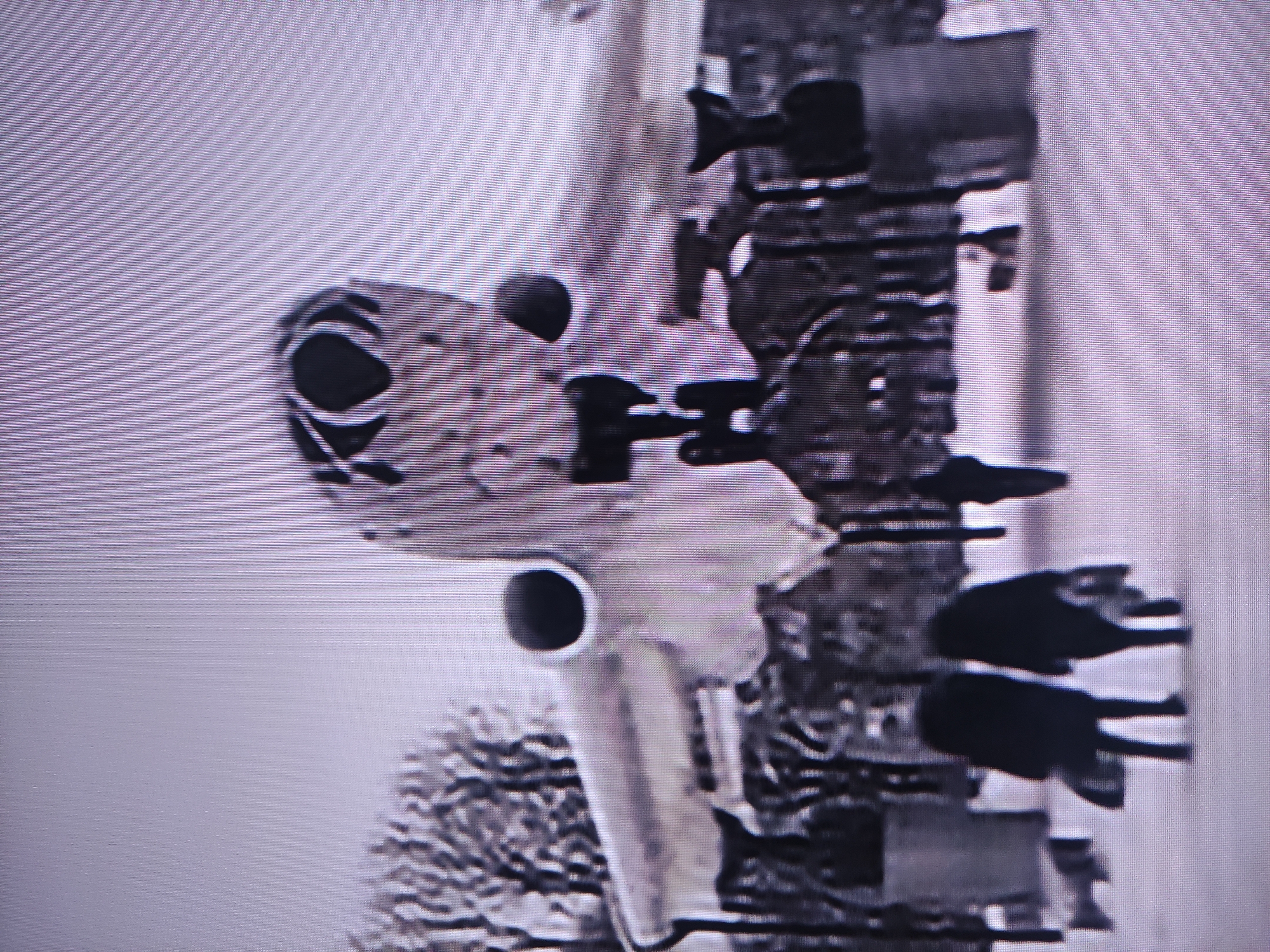 The image displays a photograph of a static model diorama featuring an aircraft. The primary subject is a light-colored model of an A-10 Thunderbolt II (Warthog) attack aircraft, oriented with its nose pointing towards the left. Distinctive features of the aircraft visible include the circular muzzle of its nose-mounted GAU-8 Avenger cannon and one of its twin turbofan engines mounted high on the fuselage.
To the right of the aircraft, a dark, textured vertical structure, resembling a tower or building, is present. Several dark, silhouetted miniature figures, likely representing soldiers or ground crew, are positioned on different levels of this structure. Some of these figures appear to be holding equipment.
In the foreground, towards the bottom left of the image, there is a blurry, light-colored, textured element that suggests a bush or tree, possibly covered in snow or frost. The background is a plain, bright, light gray or white expanse, indicative of an overcast sky or a simple studio backdrop for the diorama.
The image itself exhibits prominent horizontal scan lines and a grainy quality, suggesting it is a photograph taken of a screen or a low-resolution digital capture. No discernible text is visible on any part of the scene. The overall activity depicted is a static display of an aircraft and accompanying figures within a diorama setting.