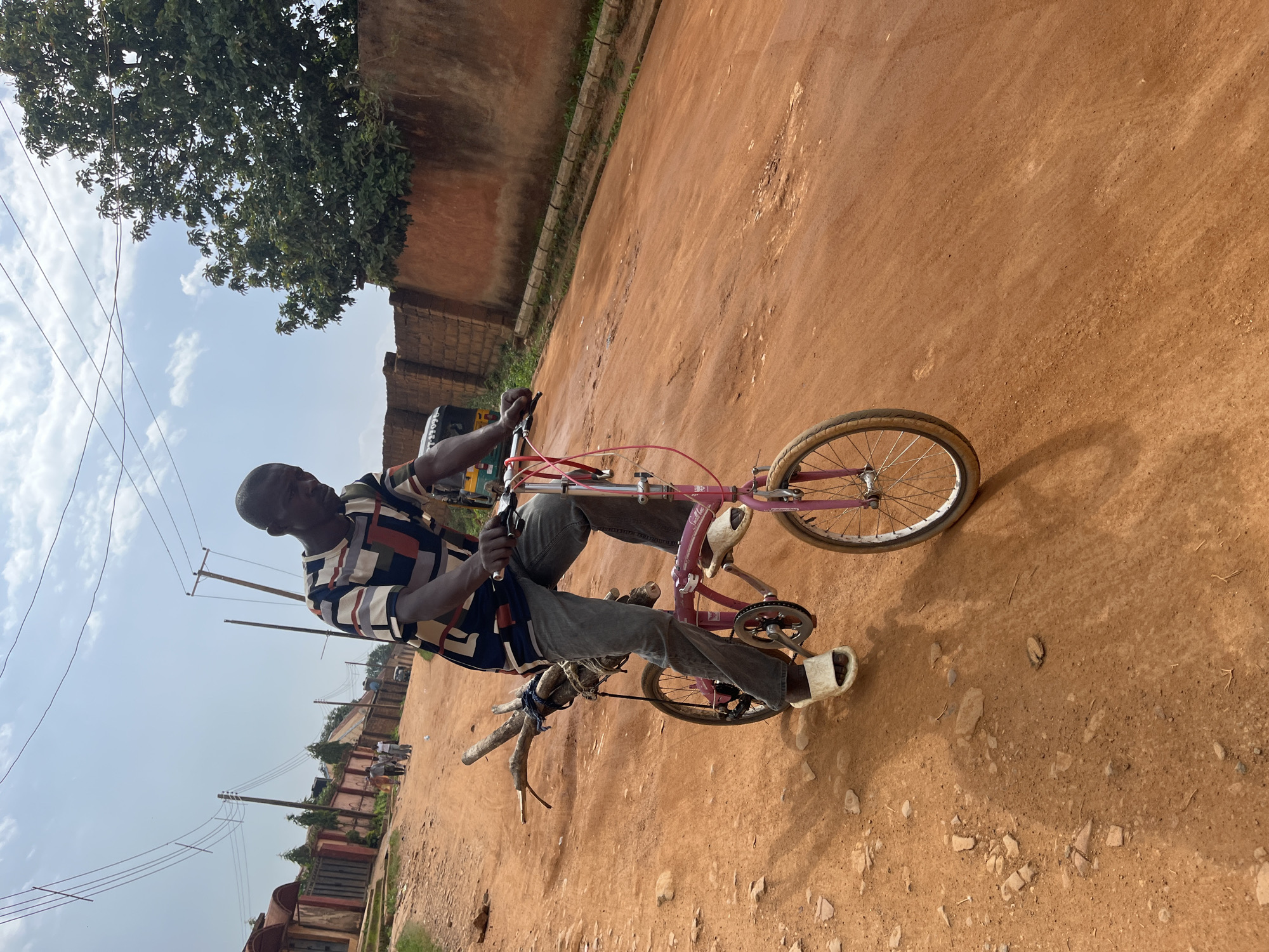An African man rides a pink folding bicycle carrying firewood on a dirt road in Barakin Akawo, Nigeria. He is wearing a striped short-sleeved shirt, grey pants, and white slide sandals. The bicycle has a small frame and the firewood is tied to the rear of the bike. The road is made of reddish-brown dirt and is uneven with scattered rocks and debris. In the background, there are buildings with reddish-brown walls, fences, trees, and utility poles with power lines. The sky is partly cloudy. Other people and vehicles are visible in the distance.