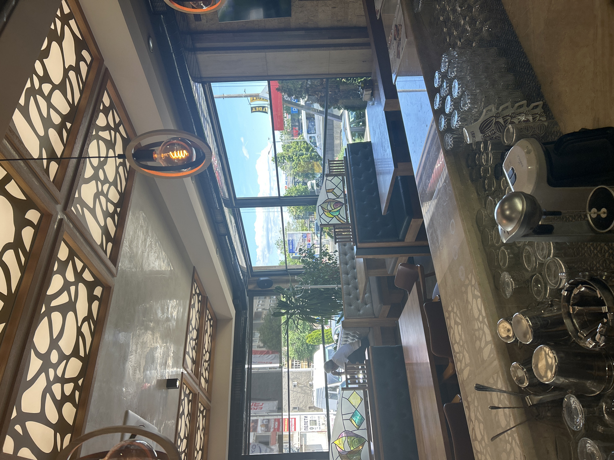 The image depicts the interior of a restaurant in Kazanlak, Bulgaria. The view is likely from a high vantage point or tilted, looking towards a window that provides a view of the street.
Architectural details include decorative wooden wall panels with a cutout pattern and modern lighting fixtures. Seating arrangements include booths and tables with chairs. The interior design has a contemporary feel.
Outside, the view shows urban elements such as buildings, vehicles, and greenery. Street signs with Cyrillic script confirm the location. The sky is blue with scattered clouds, indicating daytime.
A person can be seen near the window outside, appearing to be cleaning or maintaining the exterior. On the bar counter, a variety of glasses are neatly lined up, along with bar equipment and accessories.