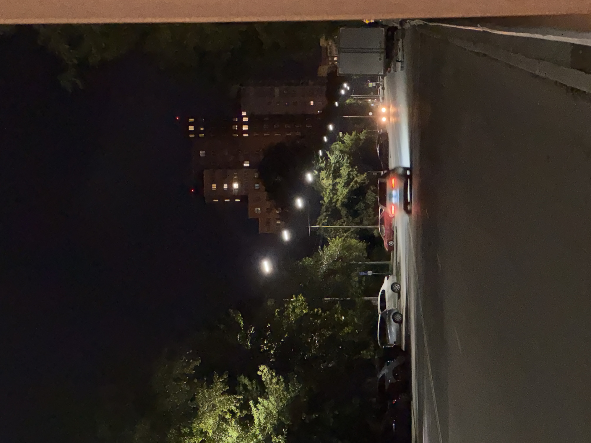 The image depicts a street scene at night in Chelyabinsk, Russia. A multi-lane road occupies the foreground and midground, with parked cars visible on the left side of the road. Several vehicles are in motion on the road, with headlights illuminating the scene. A white car is parked on the right side of the road, and a large truck is parked further down on the right. Trees line the left side of the street, their leaves illuminated by streetlights, which are also visible overhead. In the background, tall buildings with illuminated windows rise against the dark night sky. The overall impression is of a typical urban street at night, with traffic, parked vehicles, and residential buildings.
