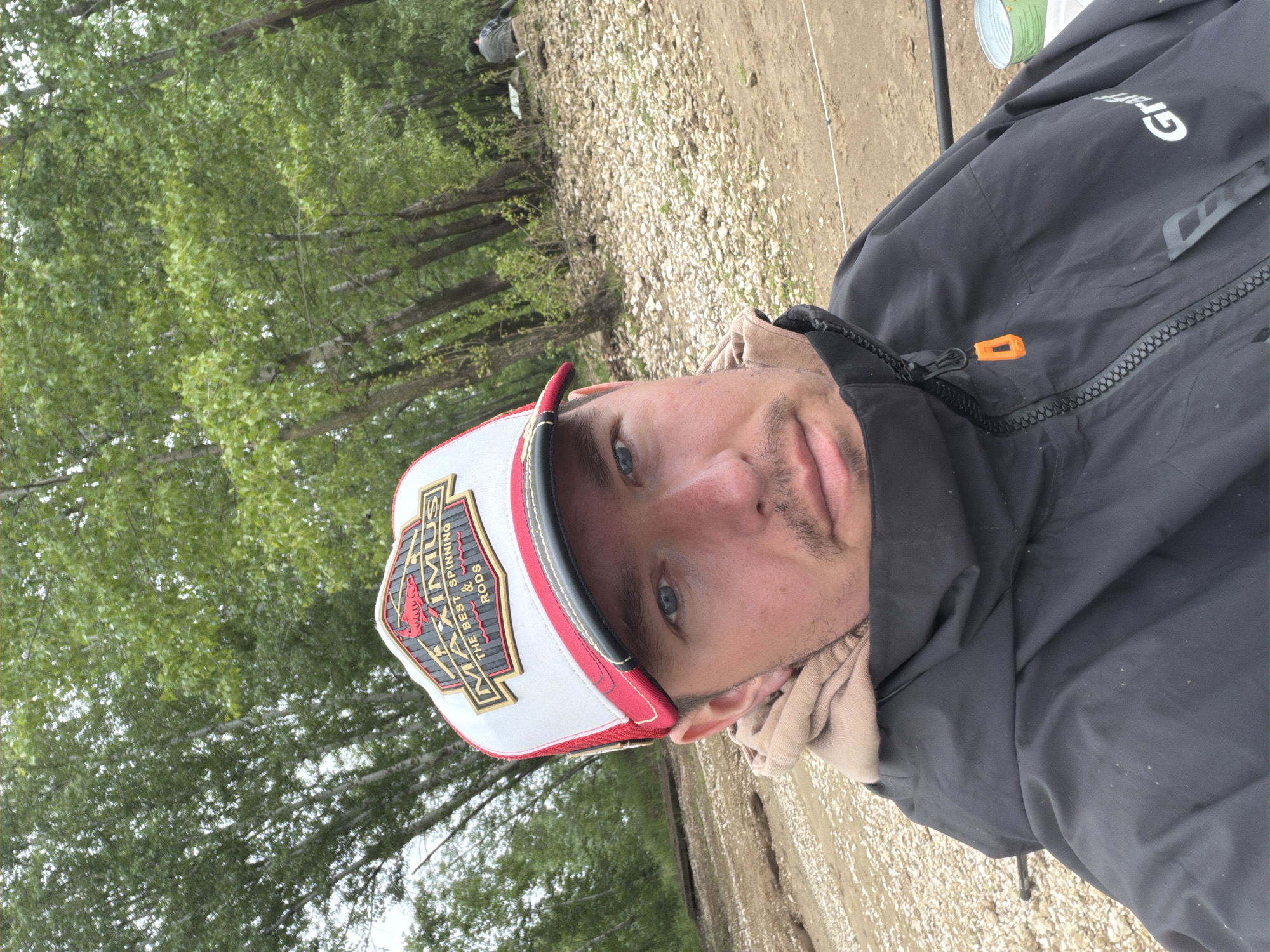 The image, oriented 90 degrees clockwise, captures a close-up portrait of a man outdoors in Noviy, Russia. The man, likely in his 20s or 30s, has light blue eyes and light stubble. He is looking directly at the viewer with a neutral expression.
He is wearing a white, red, and black trucker-style cap. The prominent patch on the cap's front features the brand "MAXIMUS" in large, stylized letters, with the slogan "THE BEST SPINNING & RODS" beneath it, accompanied by a graphic of a fish. He also wears a dark (likely black) jacket with an orange zipper pull tab. The brand name "Graf" is visible in white text on the right shoulder area of his jacket. A light brown or beige layer of clothing, possibly a hooded sweatshirt or scarf, is visible around his neck.
The setting appears to be a natural outdoor environment, possibly a riverbank or lakeside. In the background, dense green foliage and tree trunks are visible, suggesting a wooded area. The ground consists of light-colored gravel and dirt. To the upper right (when upright), another person is partially visible in the distance, closer to the ground, and a small green container or can can be seen. The presence of fishing-related branding on the cap ("SPINNING & RODS") and what appears to be a fishing line extending from the right edge of the frame suggests the man is engaged in or preparing for fishing.
The lighting indicates daytime, with an even, possibly overcast, sky as there are no harsh shadows.