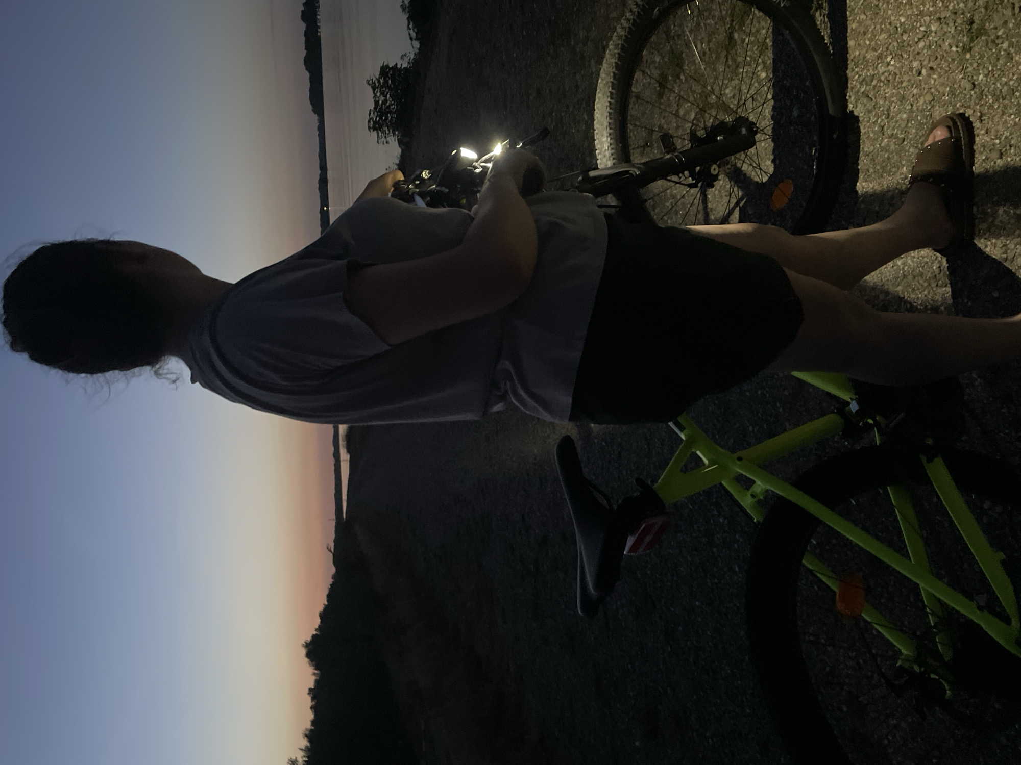A person stands next to a bright green bicycle on a gravel path at dusk. The person is viewed from the back and is looking out towards a body of water. The sky is a gradient of blue and pink, indicating sunset or sunrise. The water appears calm, with the distant shoreline visible. The bicycle has a lit headlight and is positioned to the right of the person. The overall scene suggests a moment of quiet contemplation during an outdoor activity. There is no specific indication of the location being in Nonnenweier, Germany.