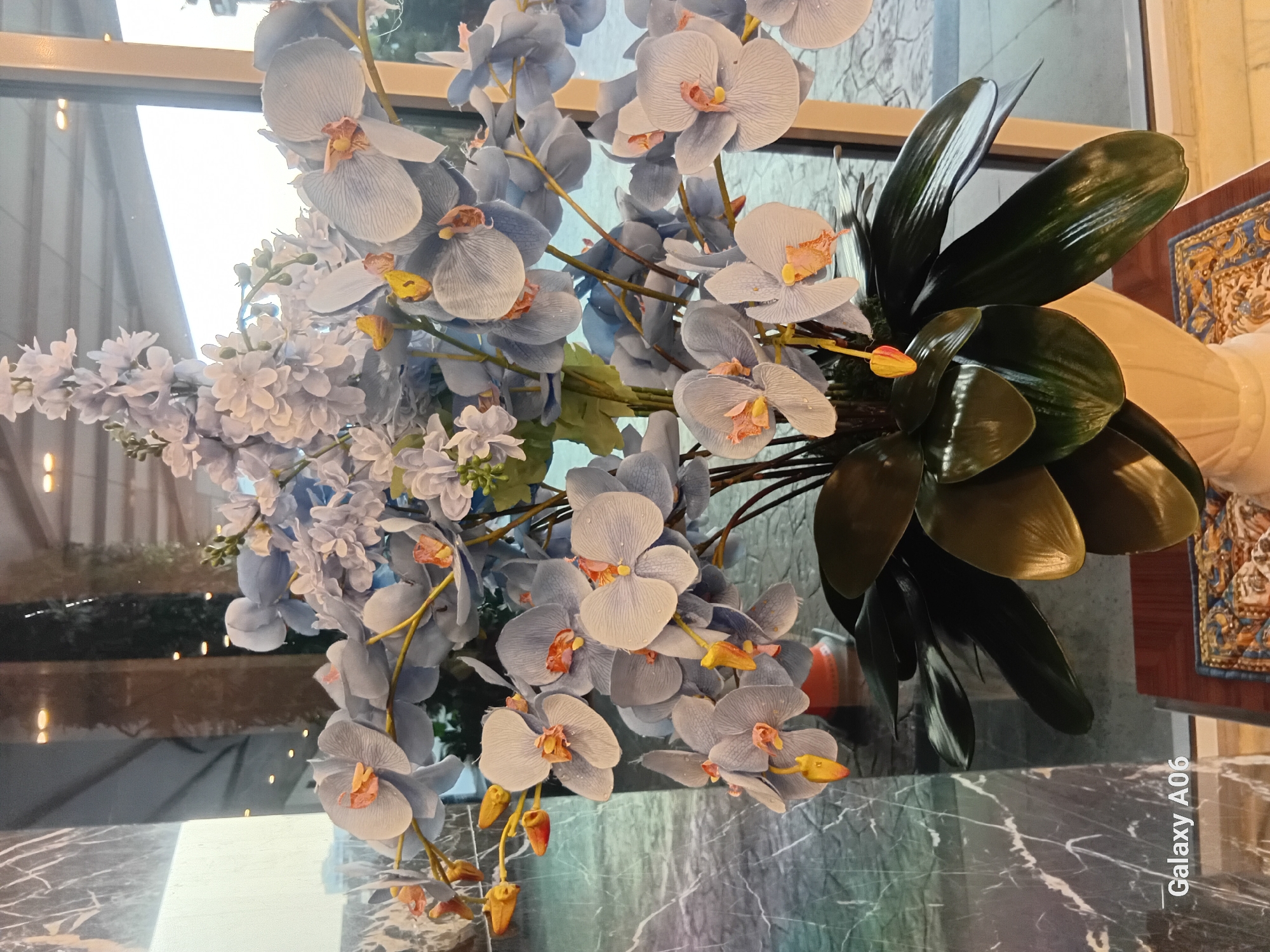 A close-up shot captures a large bouquet of artificial light blue orchids, some with pink centers, arranged in a white vase. The orchids are interspersed with smaller, lighter blue flowers and green leaves. The background reveals a reflection in what appears to be a large glass window or door. The reflection shows a ceiling with circular light fixtures, vertical columns, and a glimpse of an outdoor area with a tiled pathway and vegetation. To the left of the bouquet, a polished dark marble column with white veining is visible. In the bottom left corner of the image, the text "Galaxy A06" is present. The scene is set indoors, likely in a commercial or hospitality setting within South Jakarta, Indonesia, given the branding.
